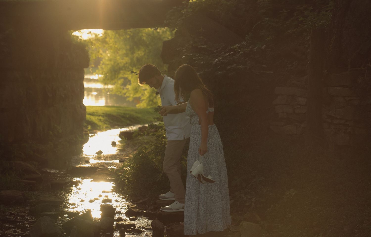 Couple standing by a sunlit stone bridge over a creek, bathed in golden hour light.