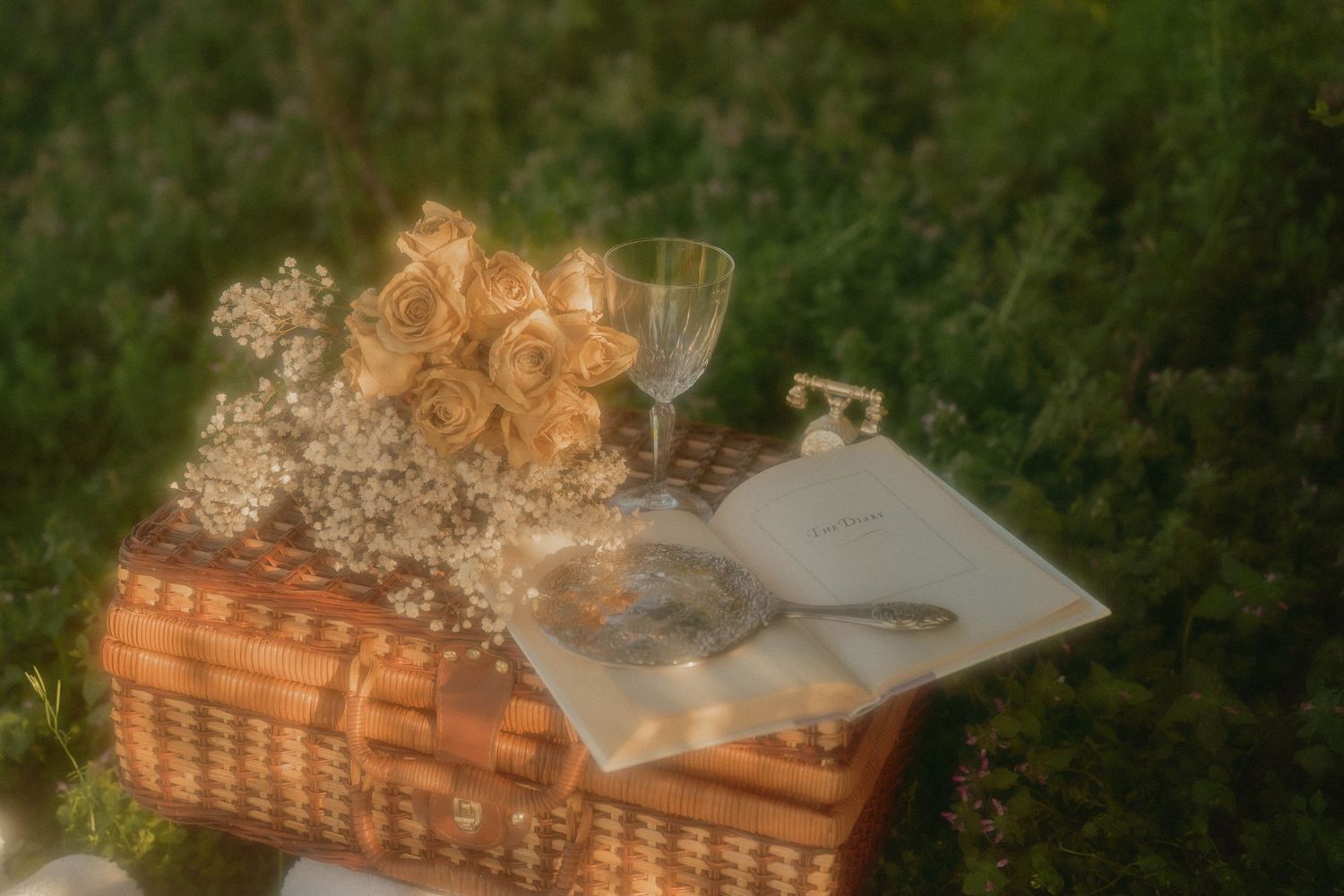 Vintage picnic basket with roses, baby's breath, open book, pocket watch, and crystal glass on green grass.
