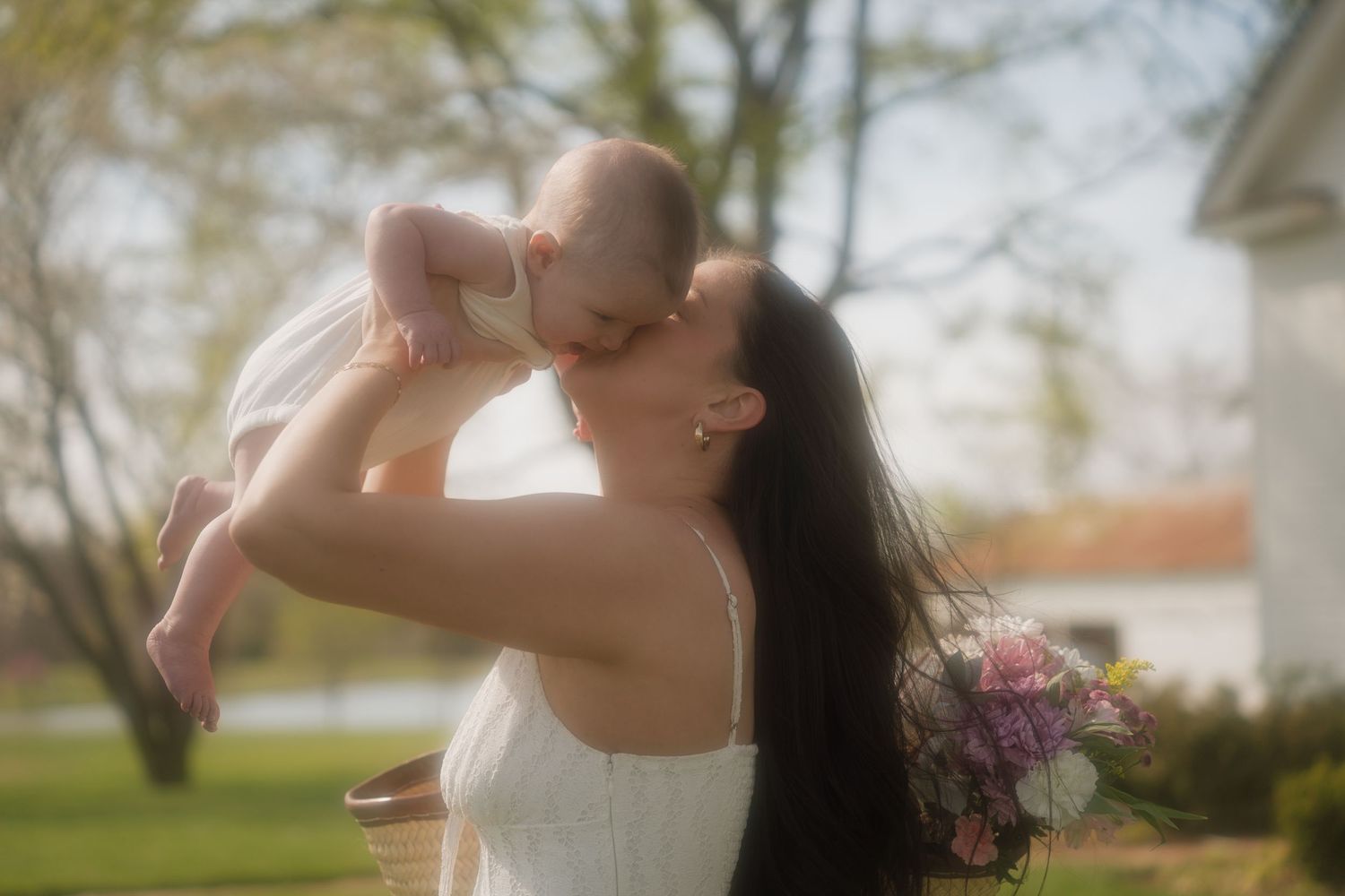 A dark-haired woman in a white dress lifts her baby outdoors, sharing a tender kiss in a sunlit garden.