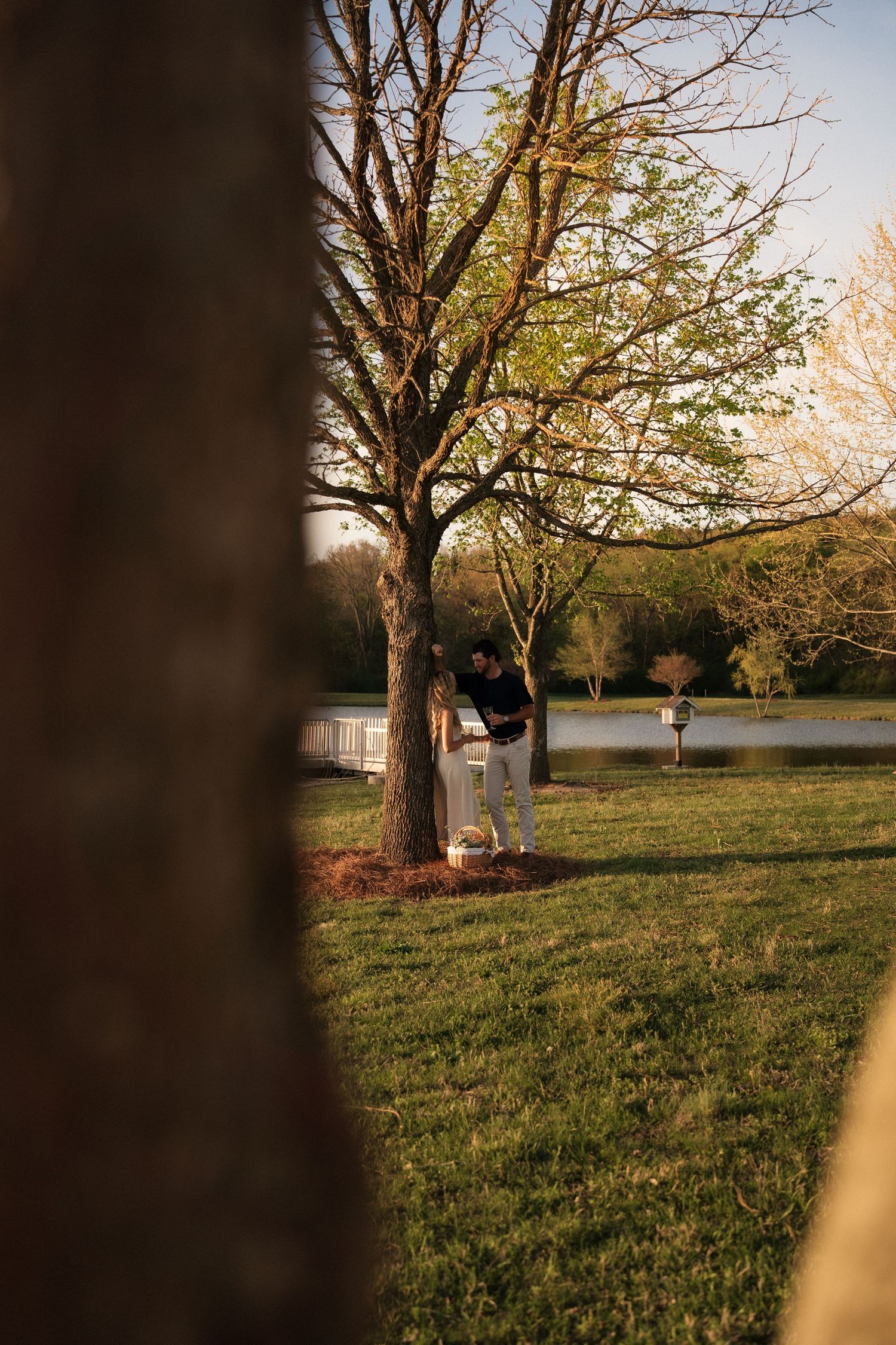 Man leaning against tree with picnic basket in golden sunset light beside tranquil pond.