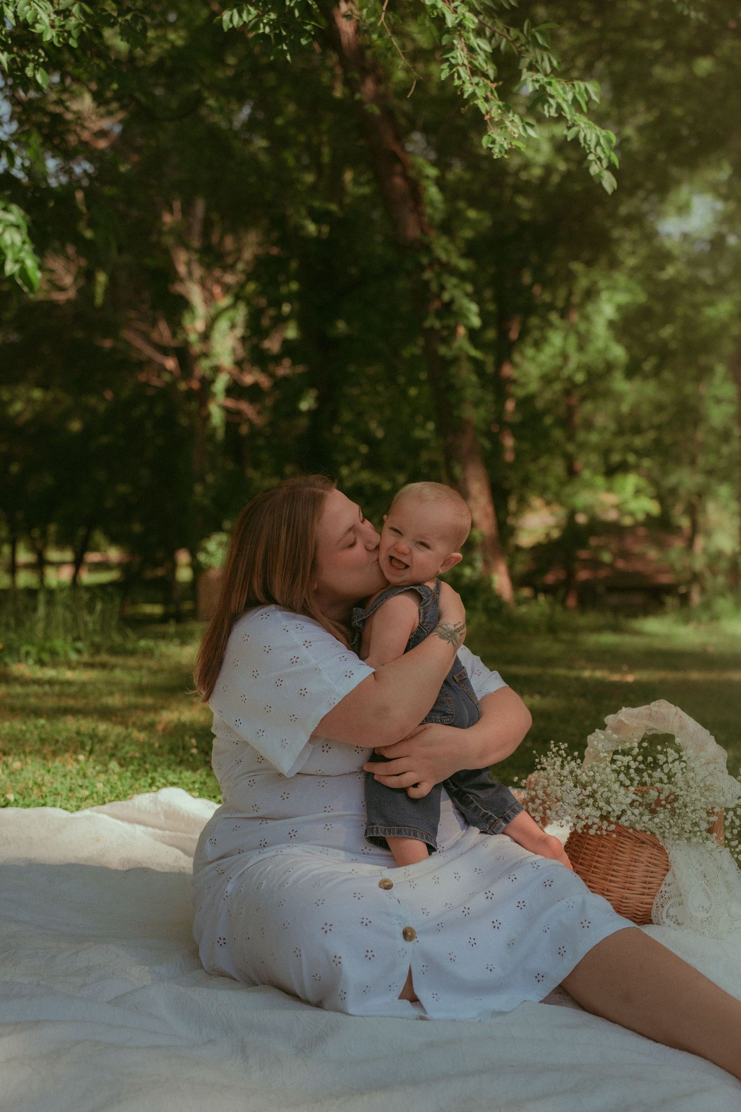 A mother kisses her smiling baby during a sunny outdoor picnic on a white blanket in a lush green park.