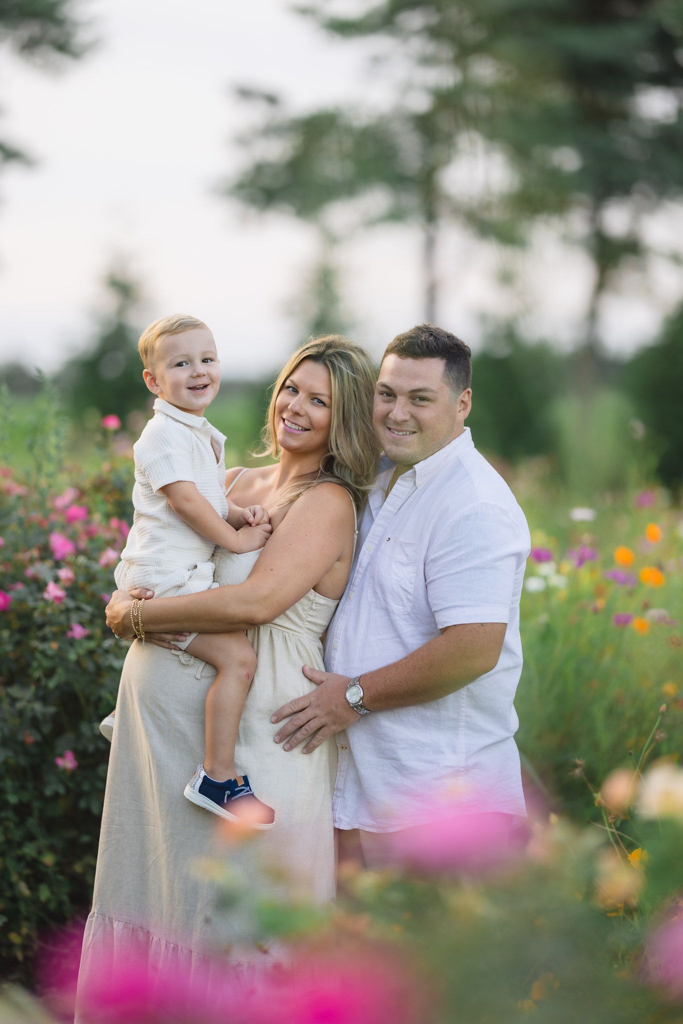 A happy family of three poses in a colorful flower field in Burlington County, New Jersey for a maternity flower field session