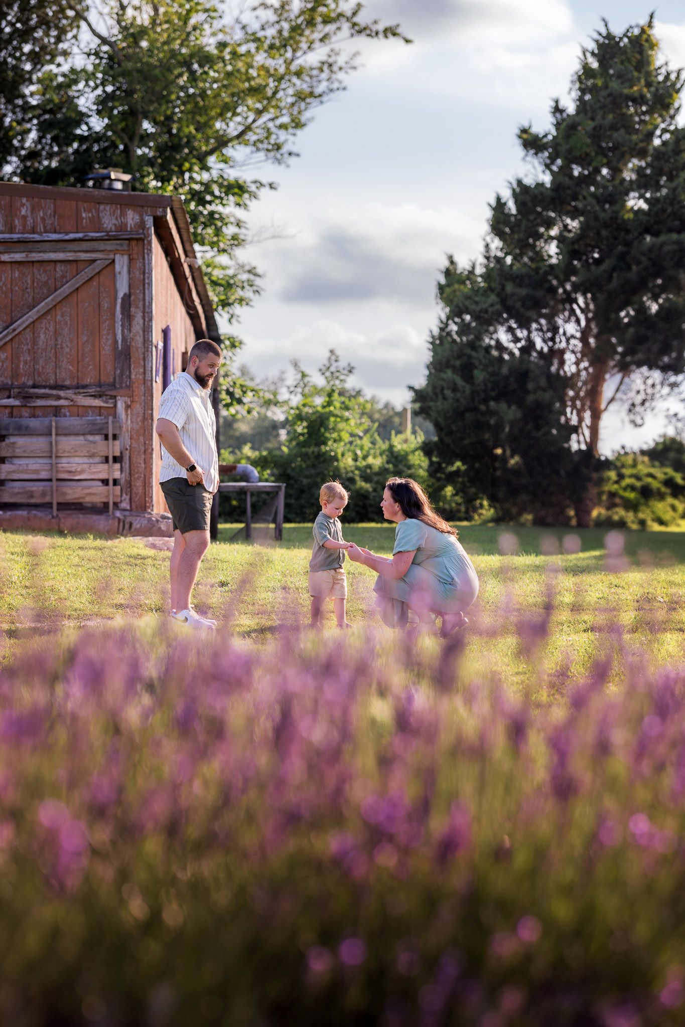 Family with young child playing in a sunny lavender field near a rustic wooden barn during a family photo session in South Jersey