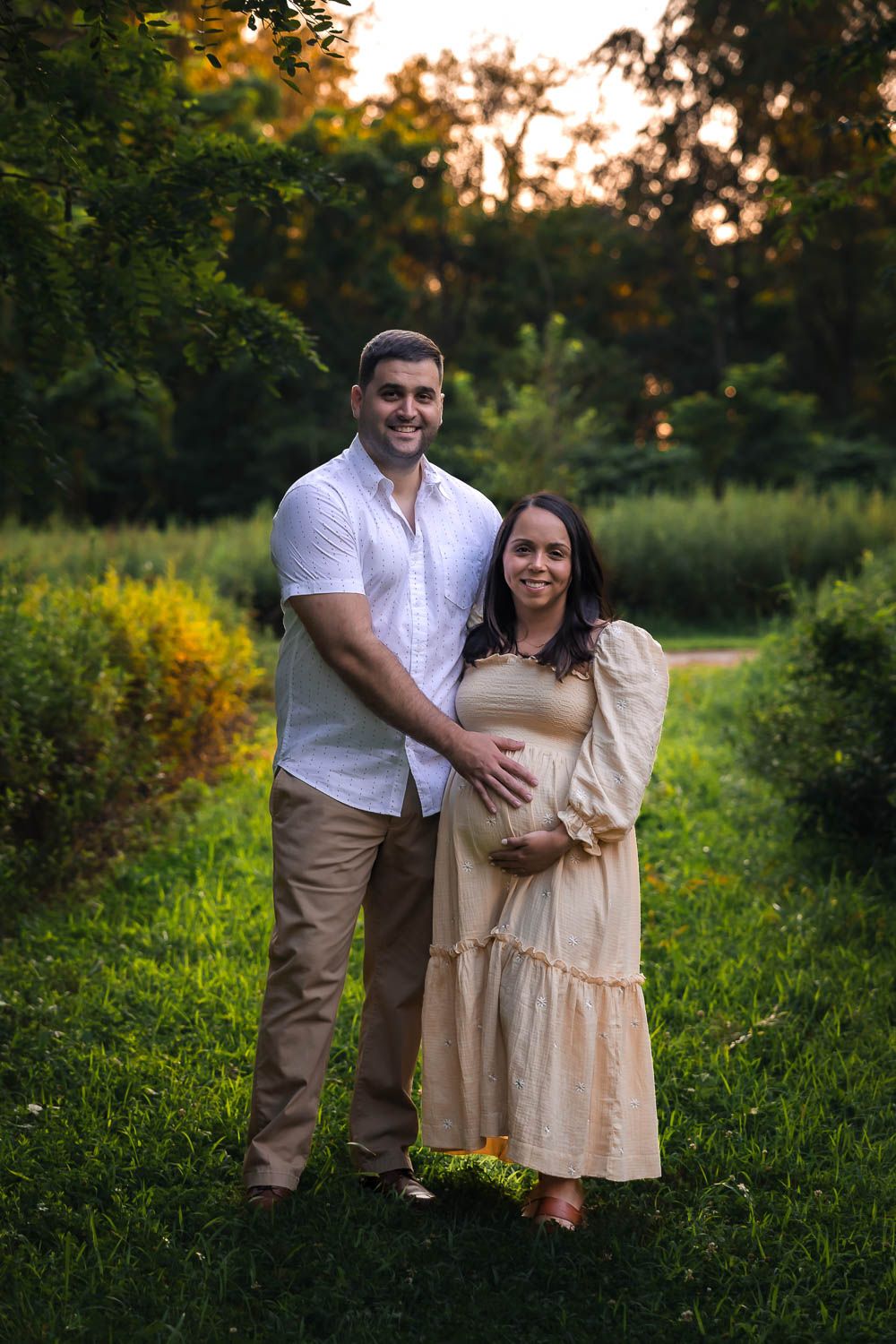 Couple posing in grassy green pathway at Palmyra Nature Cove in Palmyra, NJ during golden hour maternity photo session