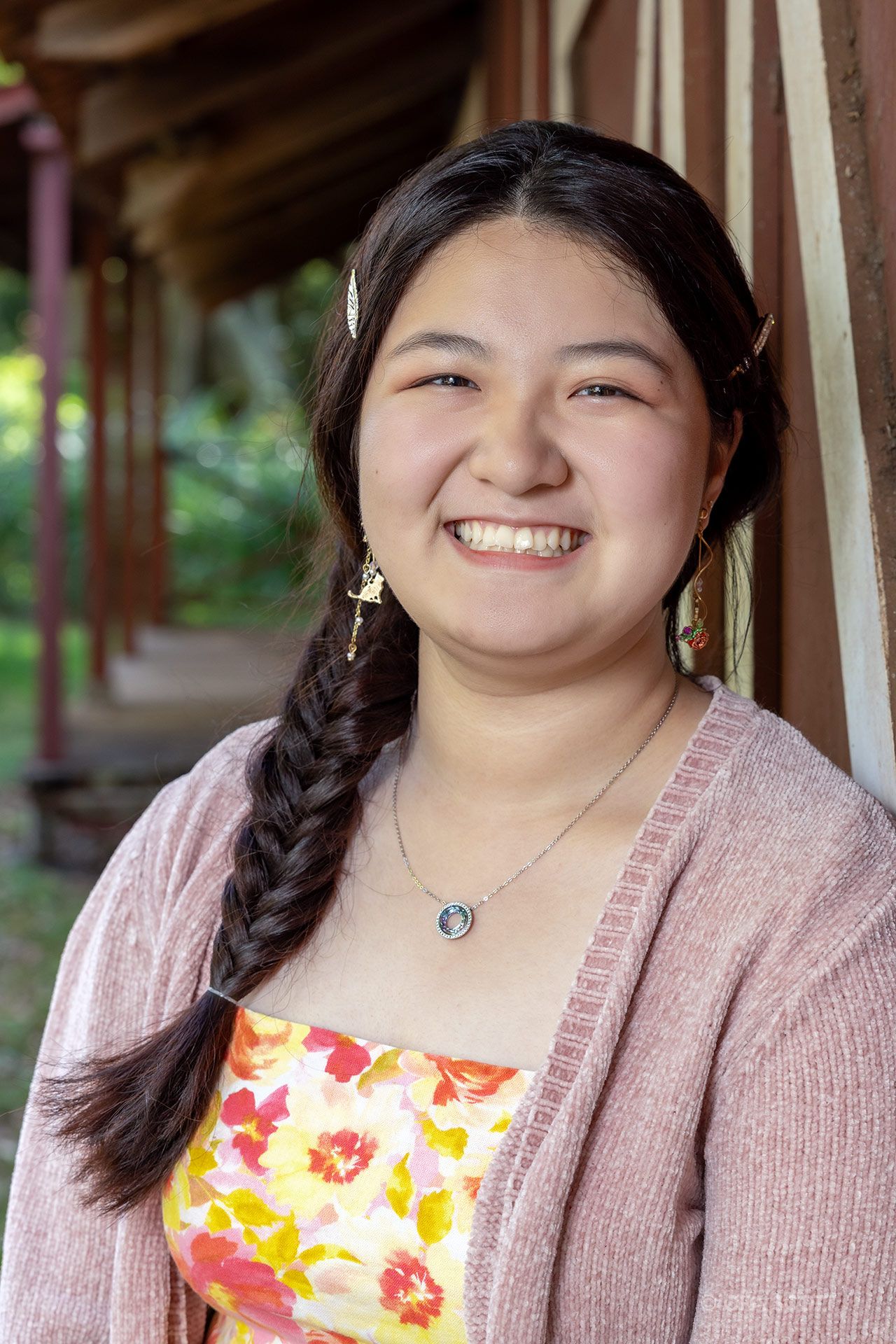 smiling girl wearing a pink cardigan and a flower summer dress having her photo taken by a red barn at her rising senior photo session