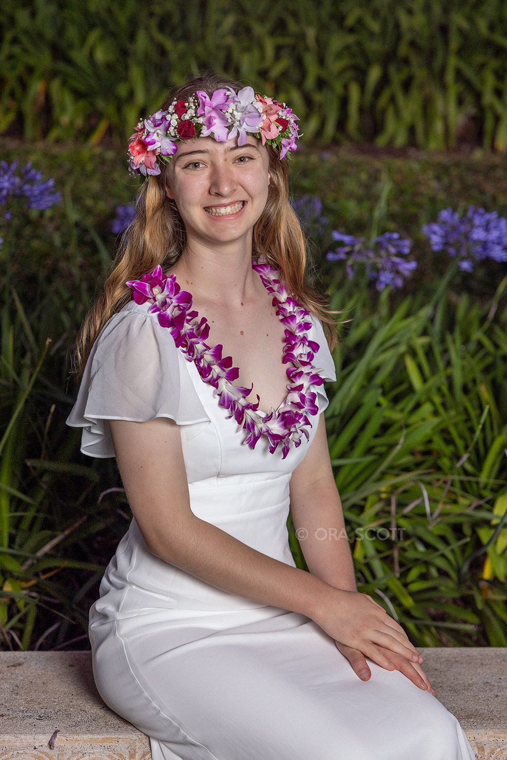 girl smiling and wearing head lei flower crown and orchid lei and a white graduation dress at her senior photo session