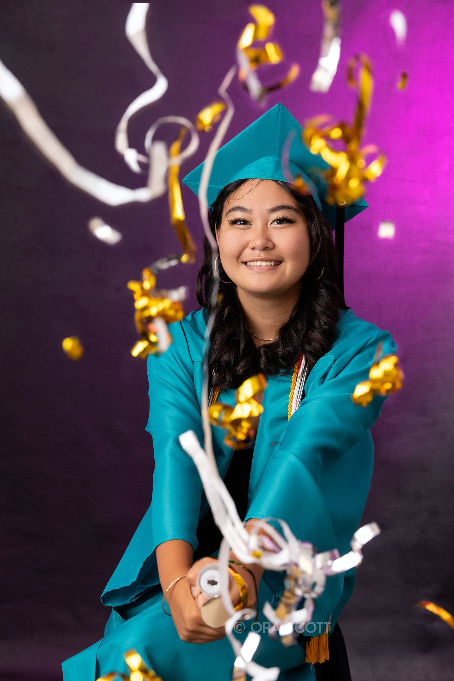 senior girl in teal graduation cap and gown using a celebration popper at her senior photo session