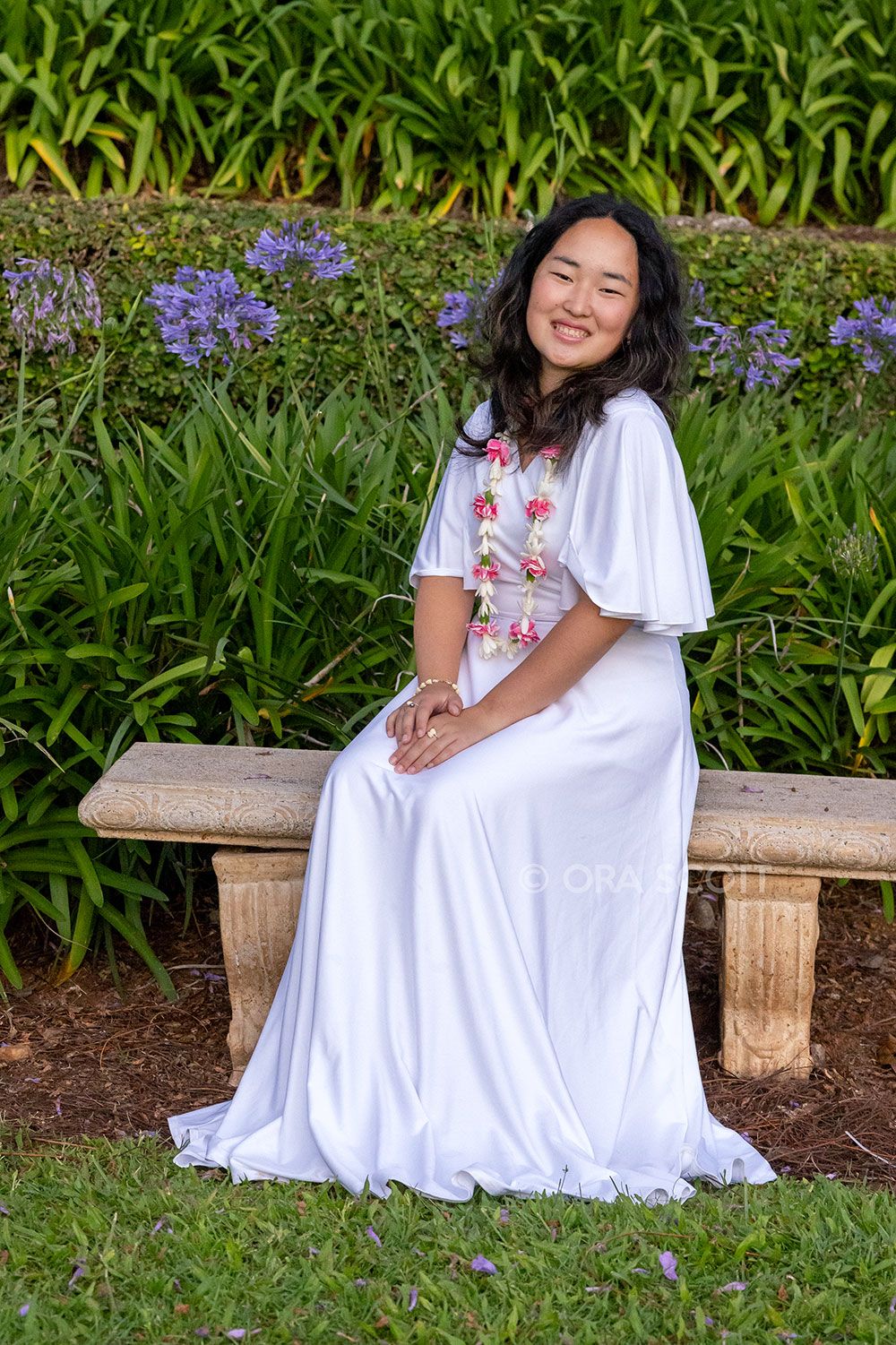 smiling girl in white dress with flower lei sitting on a bench at Seabury Hall at her senior photo session