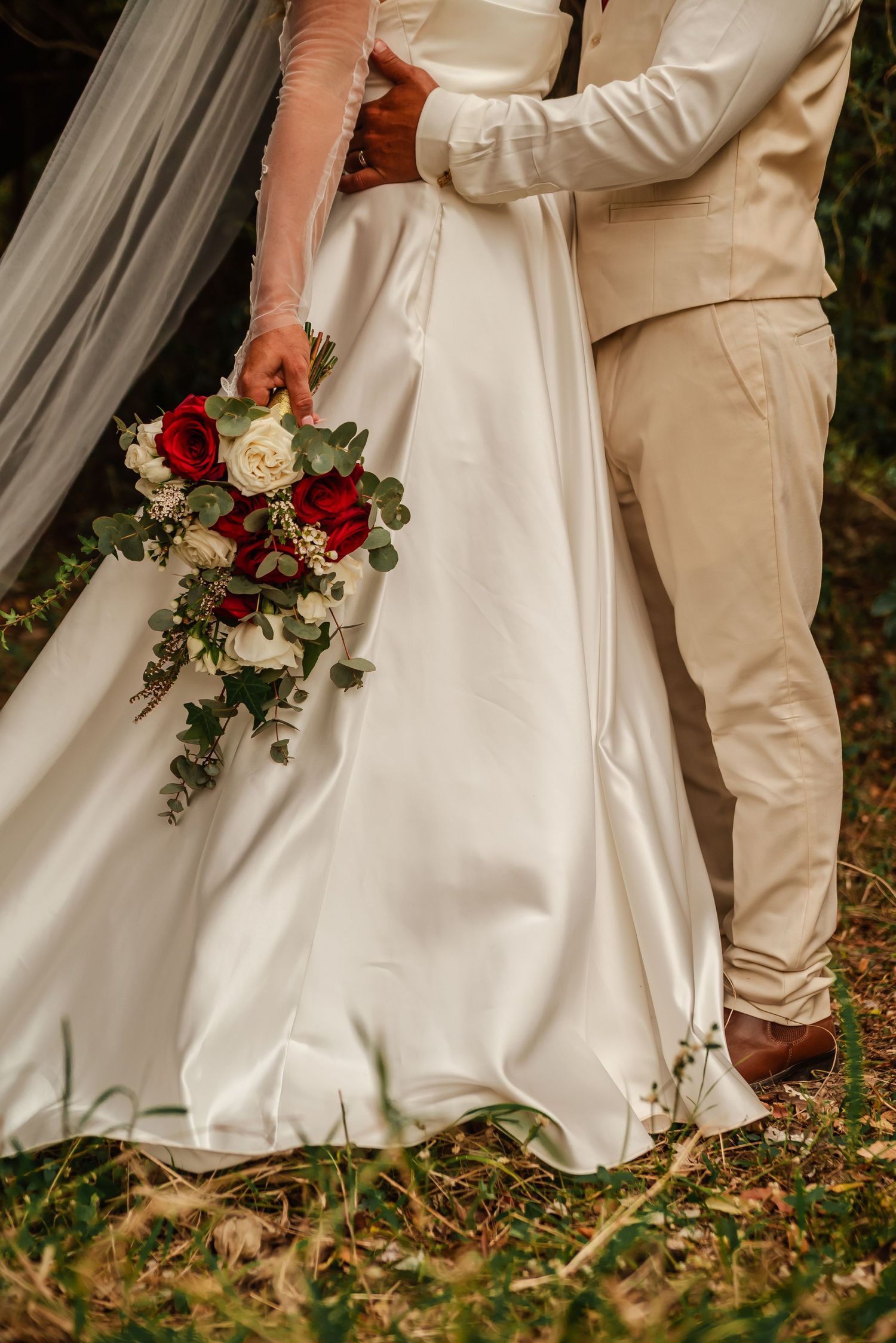 Close-up details of a wedding couple's feet and dress hem on autumn leaves with red and white floral bouquet.