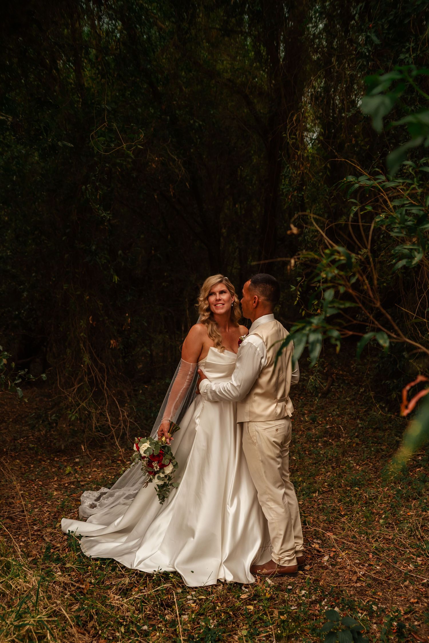 A romantic wedding couple sits together on fallen autumn leaves in a wooded forest setting.