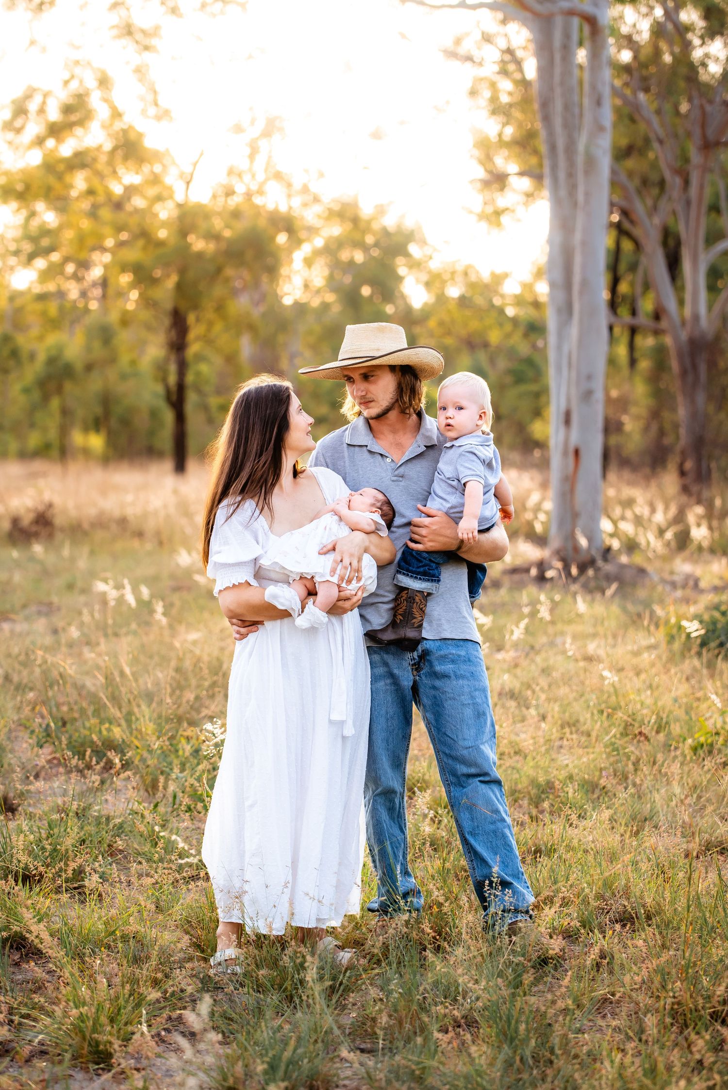 Sweet family moment captured in a rustic outdoor setting with tall trees and golden sunlight during sunset.
