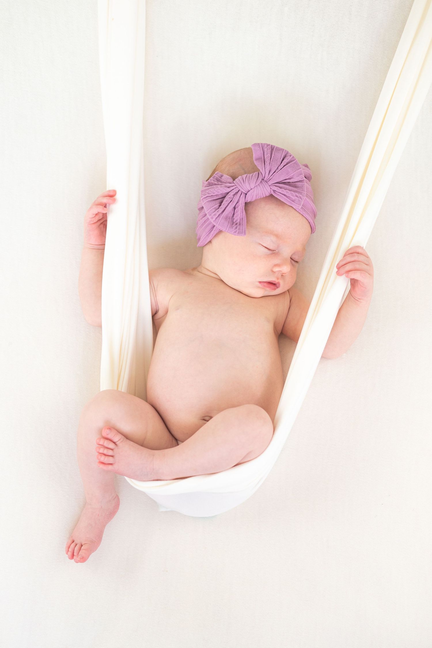 A sleeping newborn wearing a purple bow headband rests peacefully in a white fabric sling.