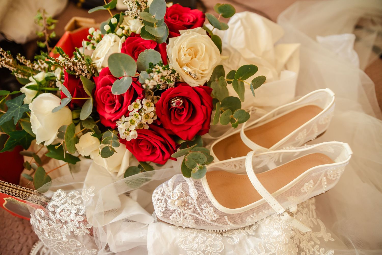 Elegant white lace bridal shoes displayed next to a bouquet of red and cream roses on delicate tulle fabric.