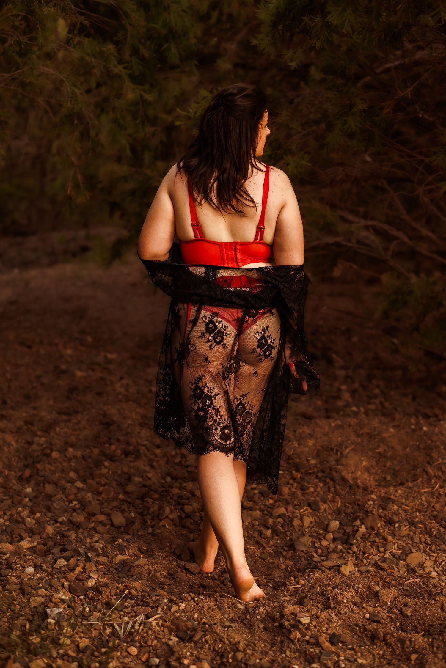 Woman in red and black lace lingerie sits on dried autumn leaves in a moody forest setting.