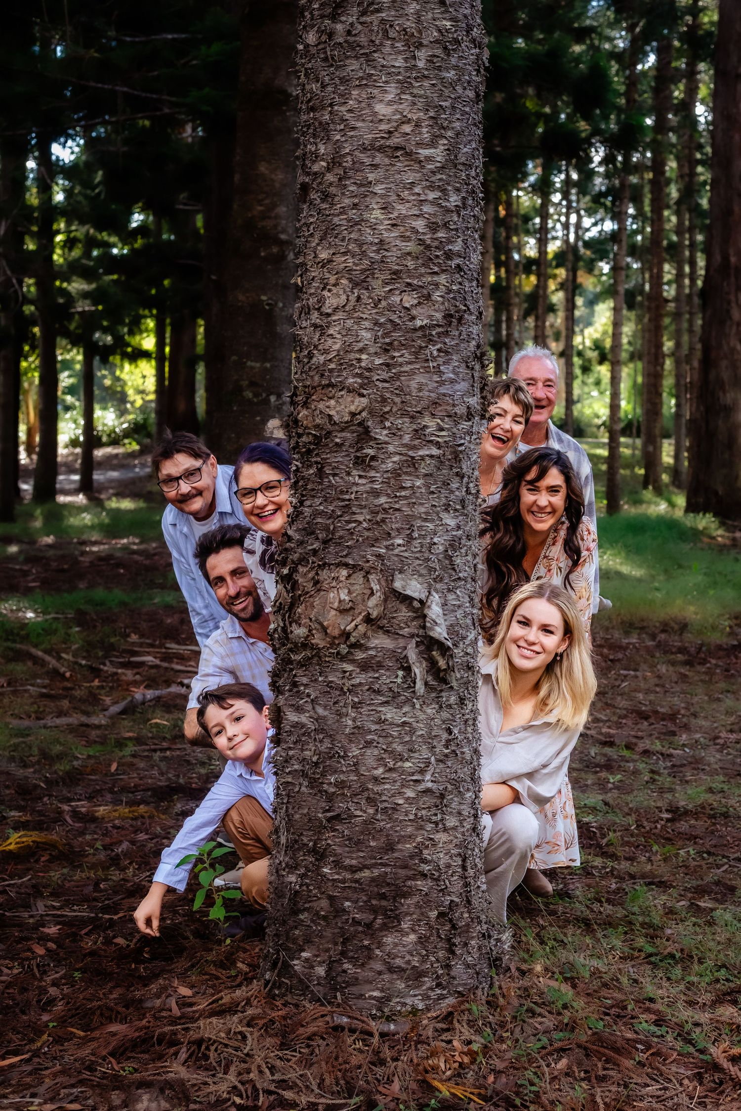 Playful group peeks from behind large tree trunk in forest during outdoor photoshoot.