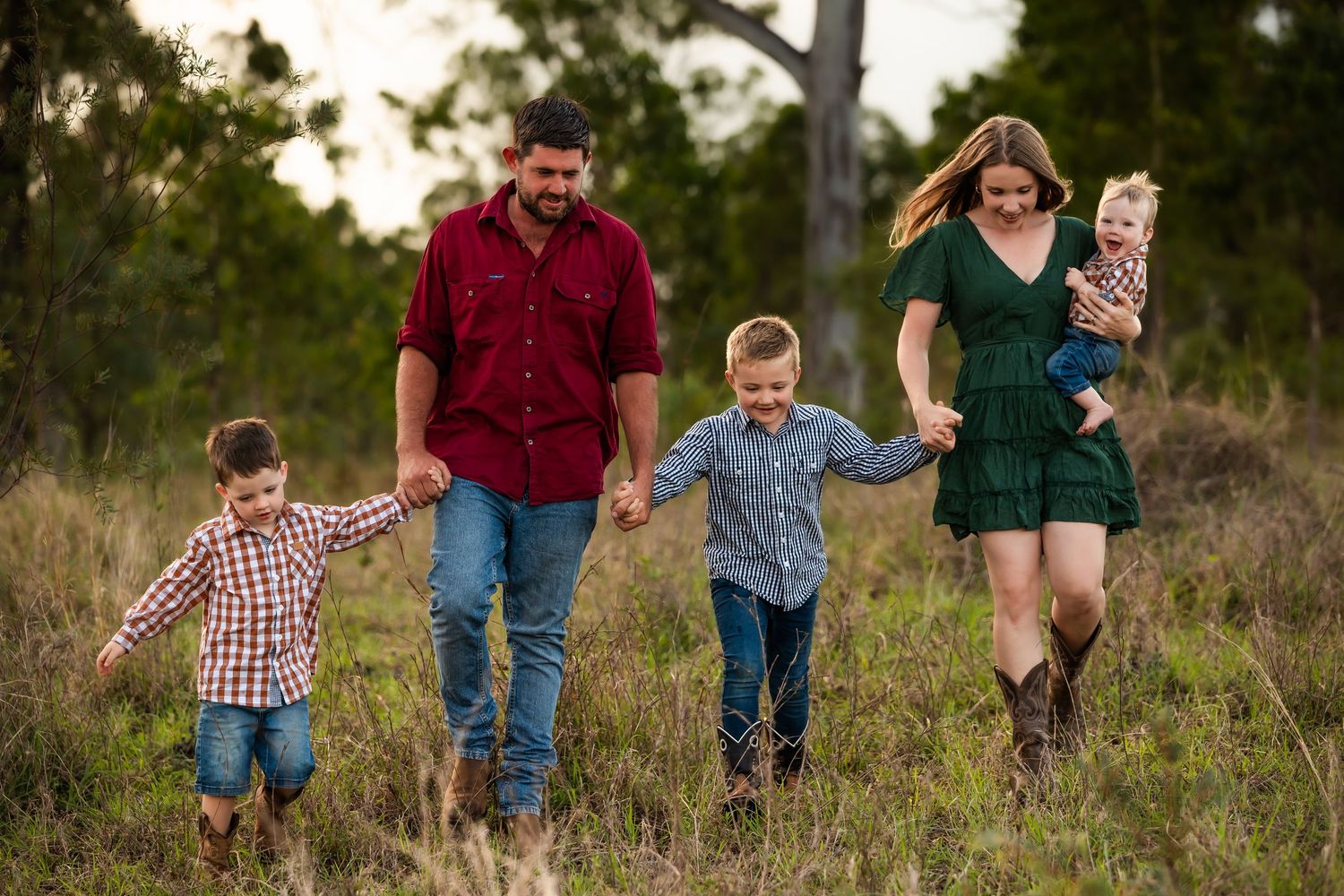 A family of four walks together holding hands through a grassy field at sunset in the countryside.