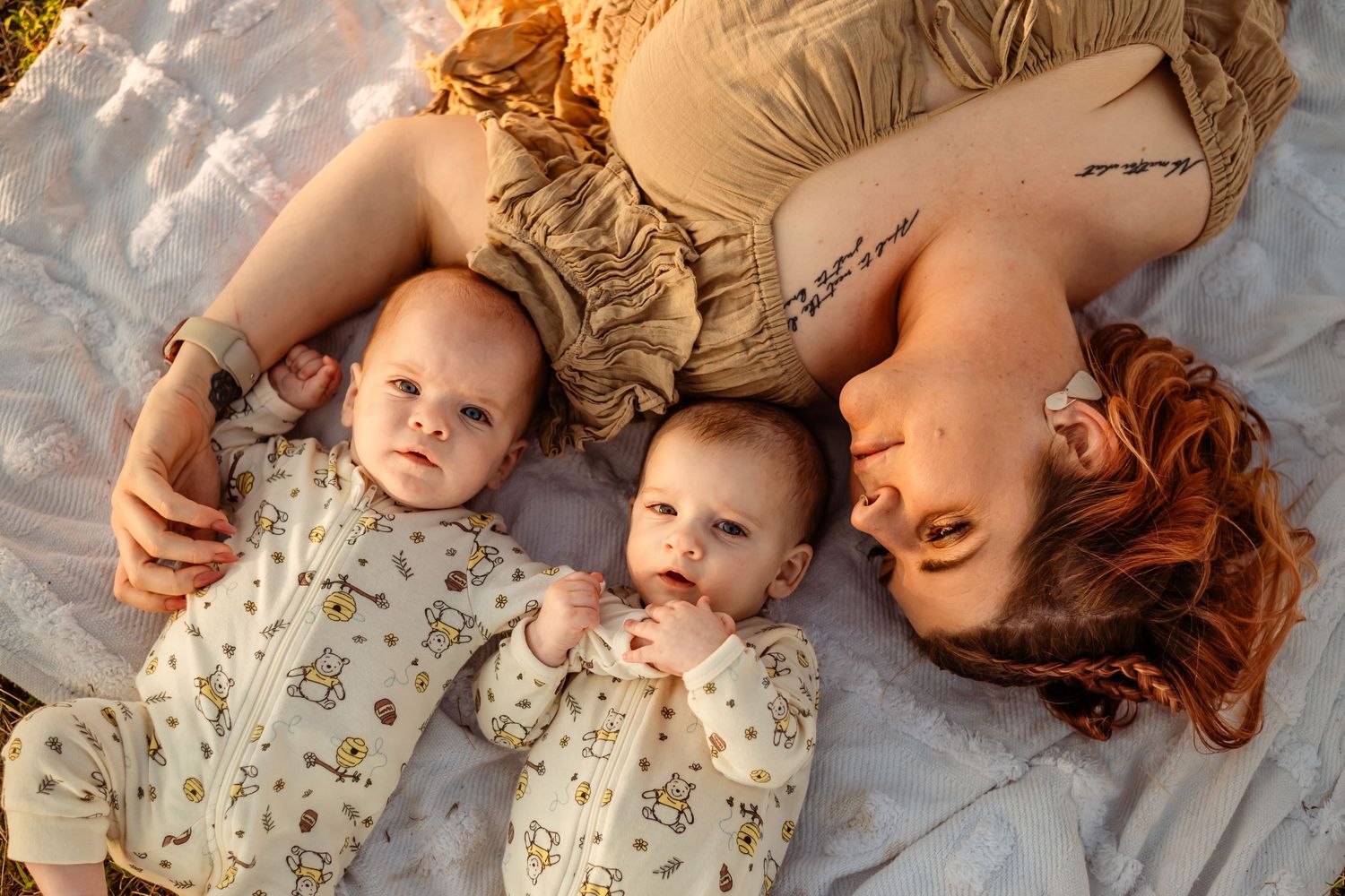 A parent and two infants wearing matching floral outfits lay together on a blanket during a golden hour photoshoot.