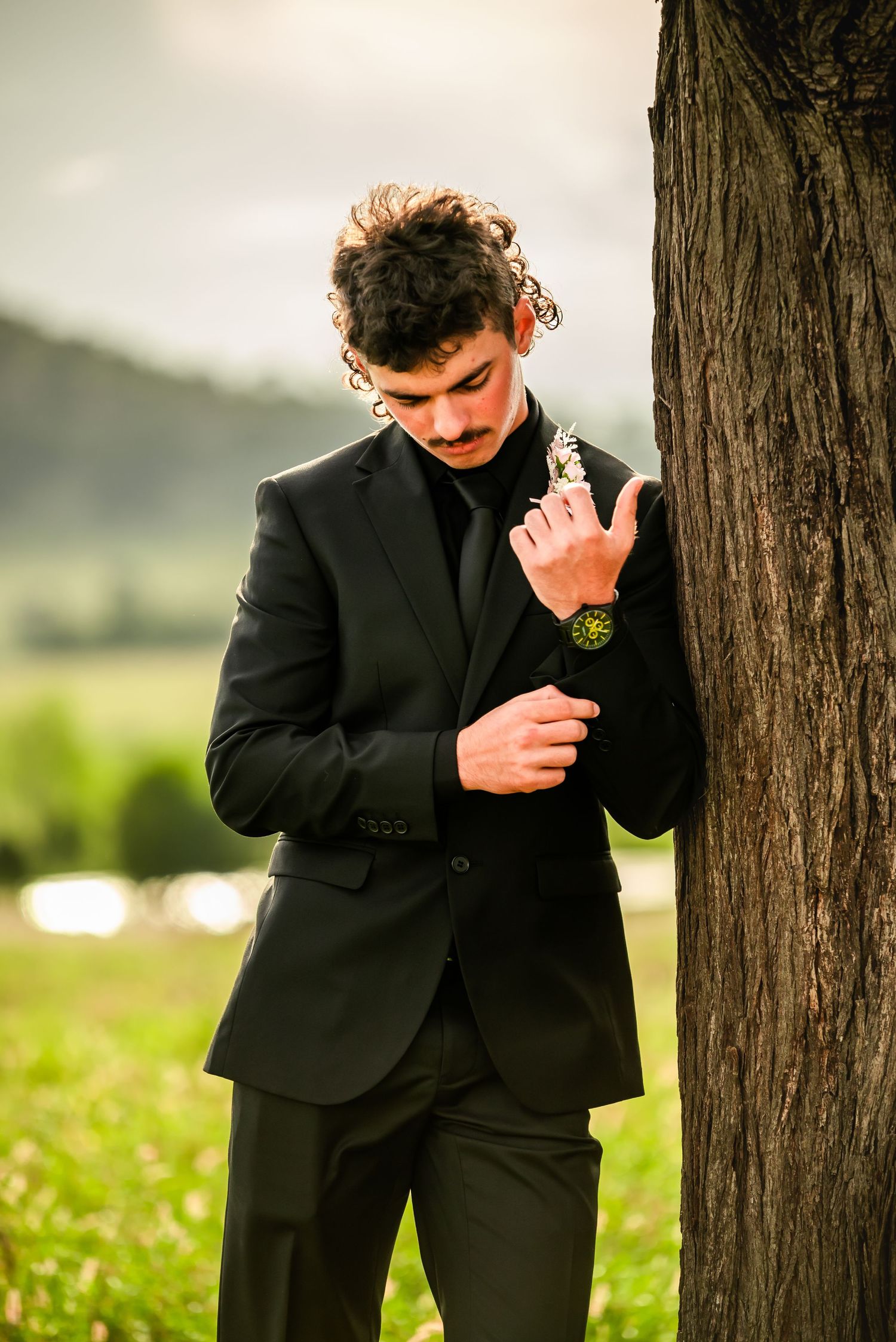A groom in a black suit leans thoughtfully against a tree trunk in an outdoor setting.