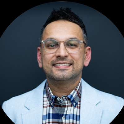 Professional headshot of someone wearing glasses, a plaid shirt and light blazer against dark background.