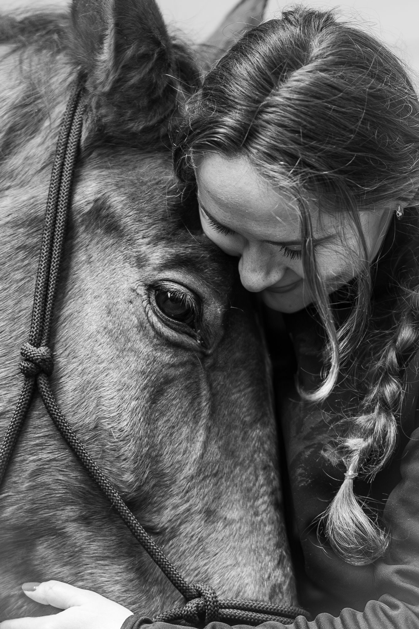 Black and white close-up of a braided young woman lovingly pressing her face against a horse.