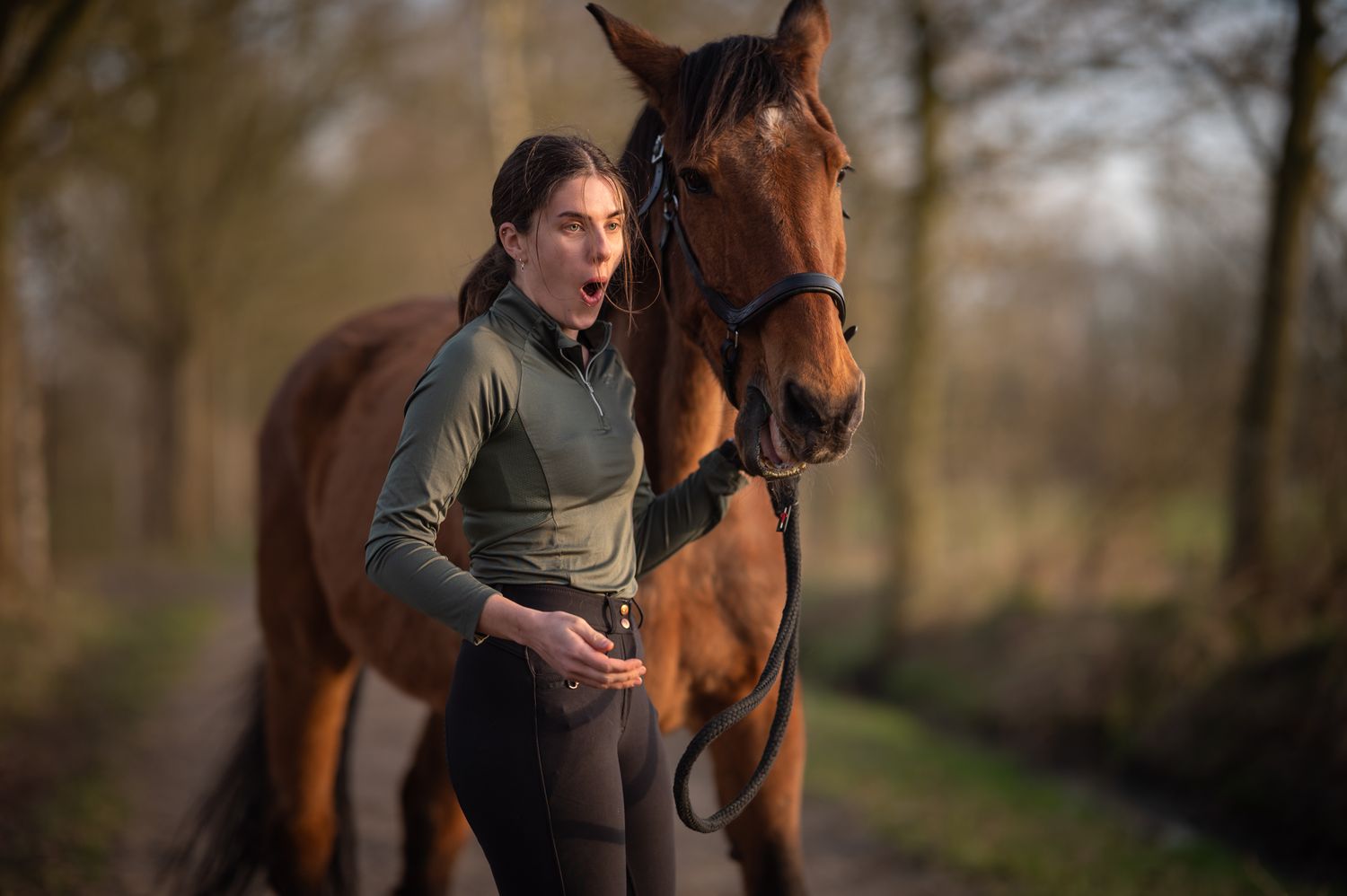 A surprised young woman standing beside a chestnut horse on a wooded forest path.
