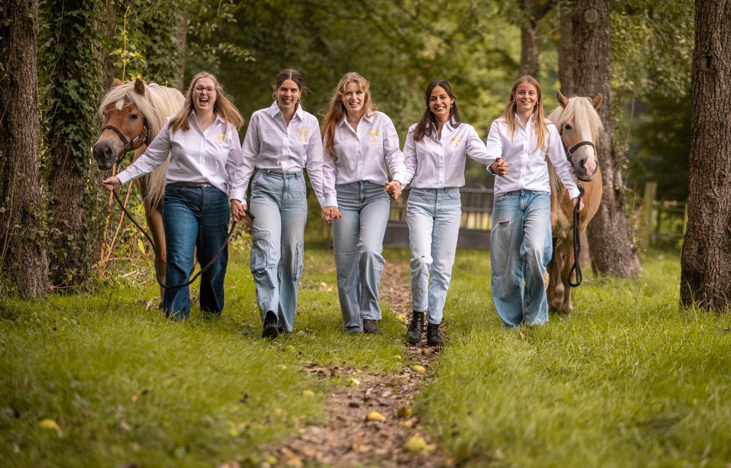 Five women in white shirts and jeans walking together with horses in a forest.
