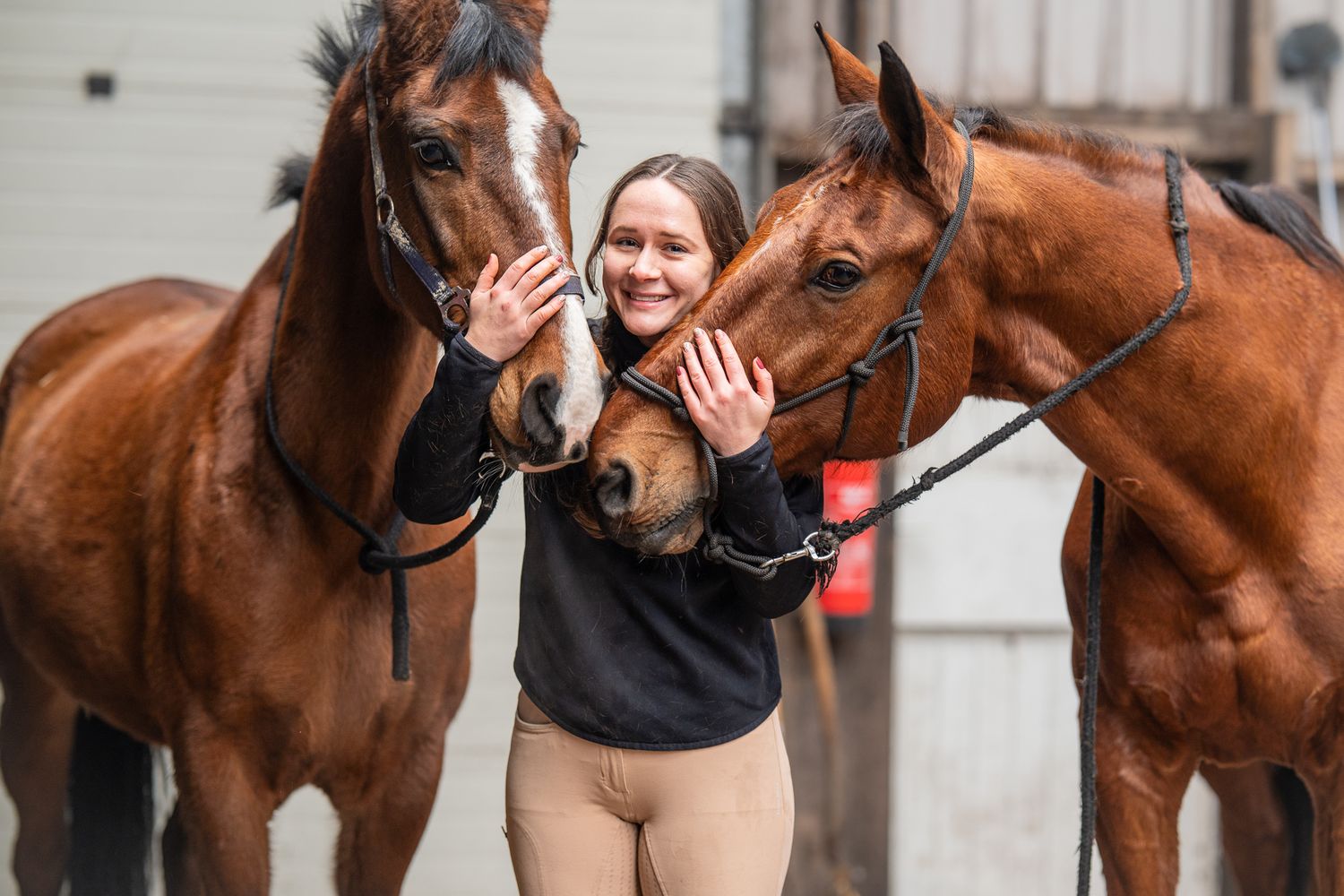 Smiling woman standing between two chestnut horses outside a stable.
