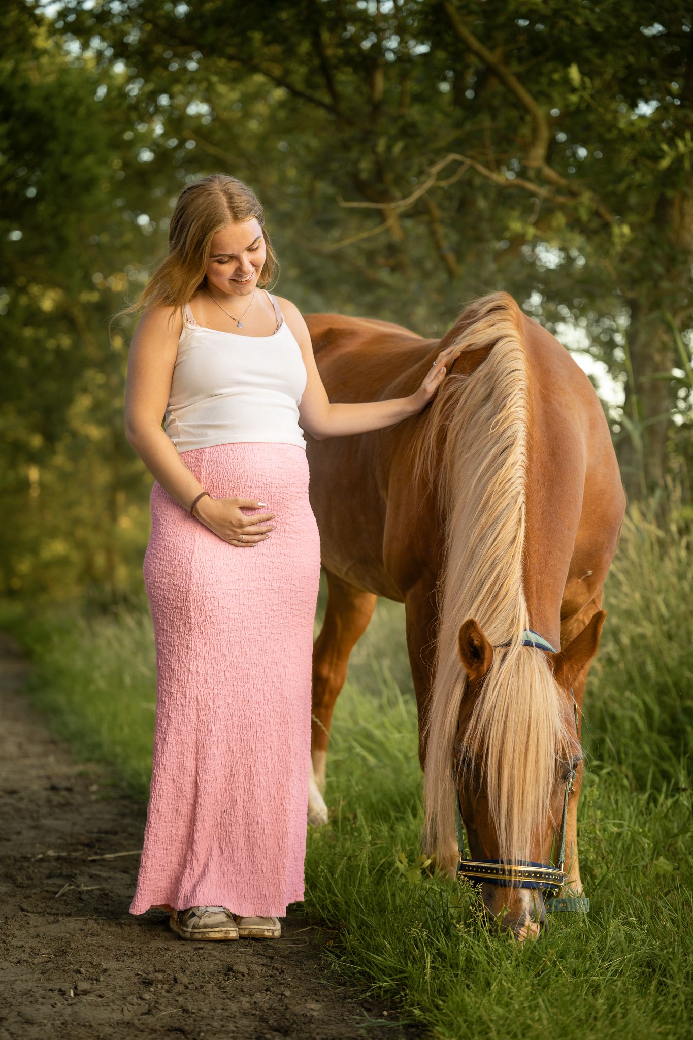 Pregnant woman in pink skirt gently petting a palomino horse on a sunny path.
