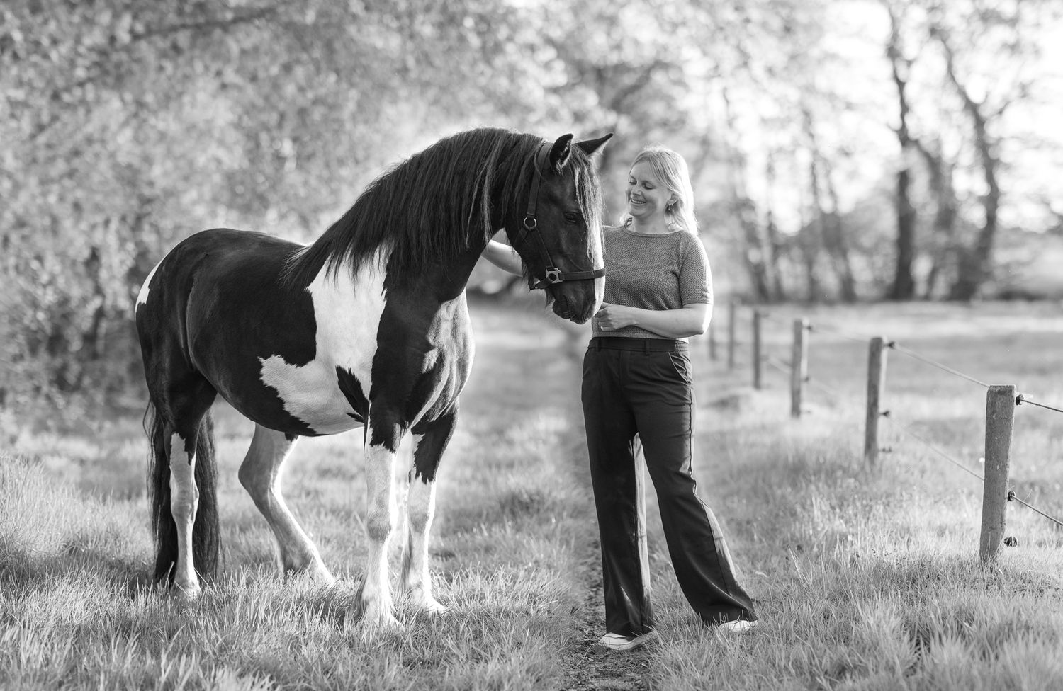 Black and white photo of a blonde woman standing beside a paint horse in a sunny field.