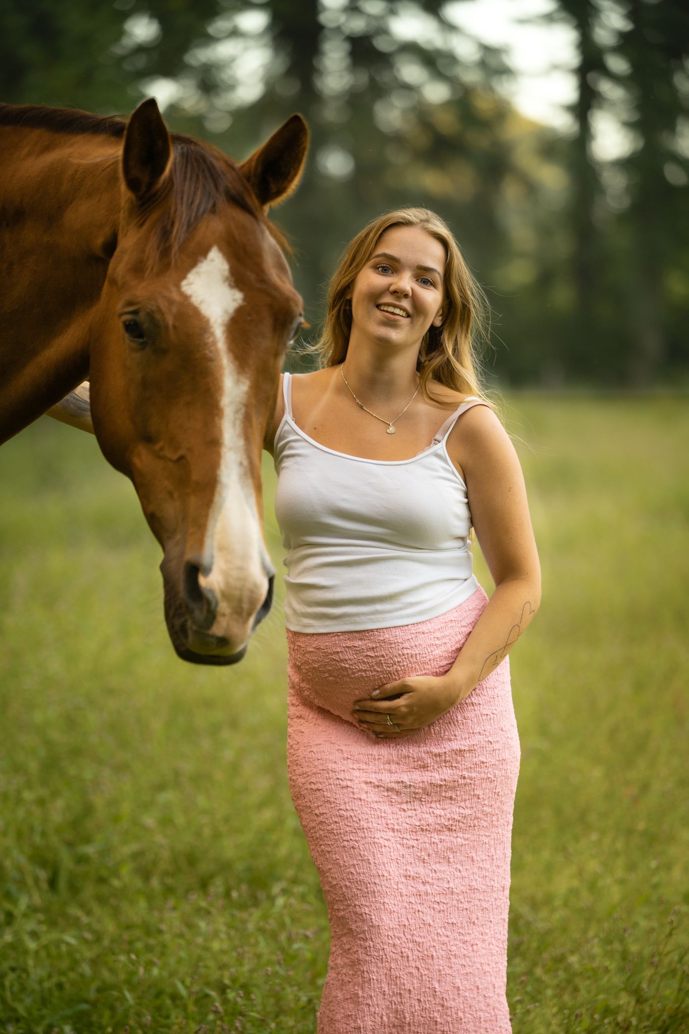 Smiling pregnant woman in pink skirt standing beside a chestnut horse in a field.