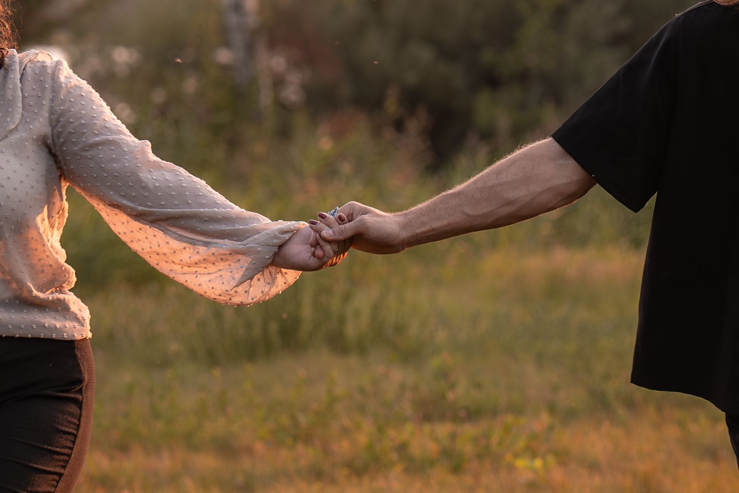 Two people holding hands in a sunlit meadow with golden hour light in background.