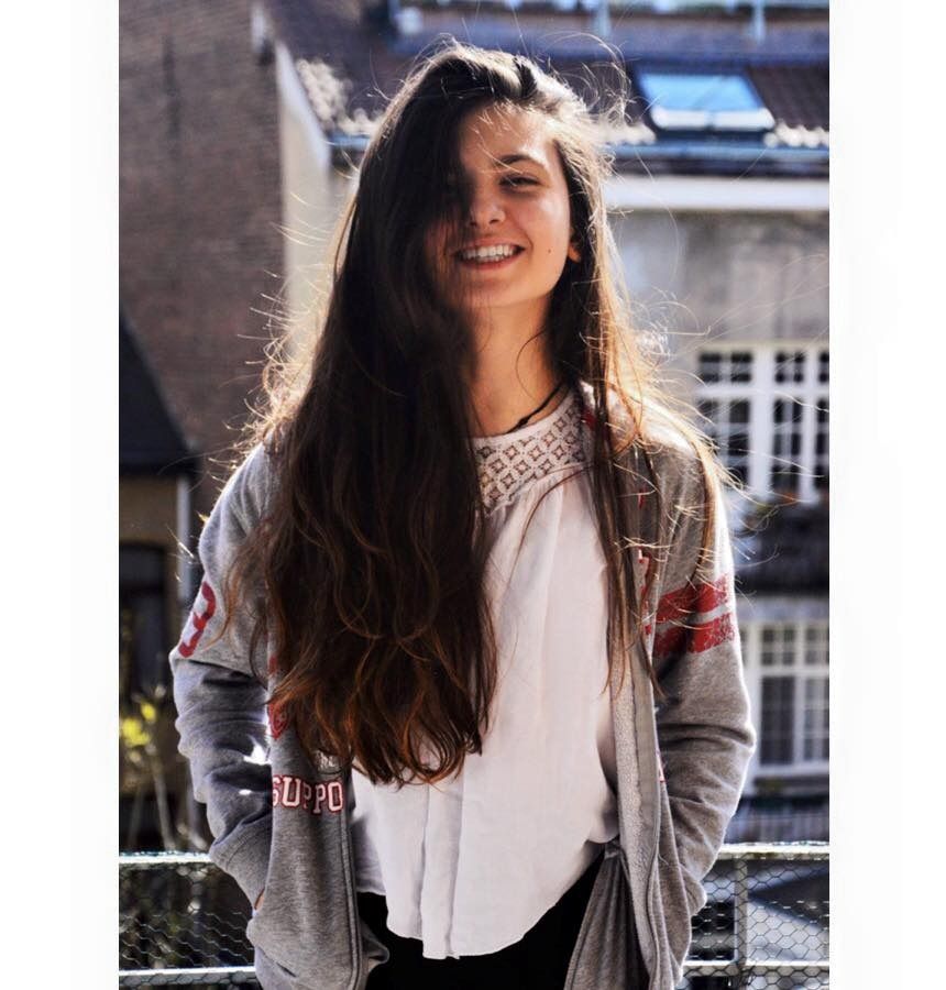 A young woman with long wavy brown hair smiles outdoors wearing a grey hoodie on a sunny day.