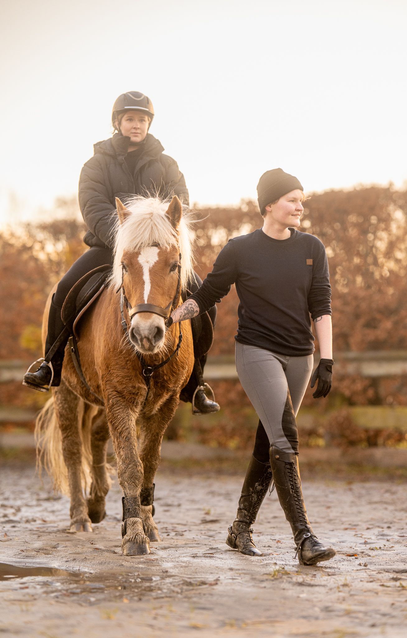 Young rider on chestnut horse being led by instructor on sandy track in golden morning light.