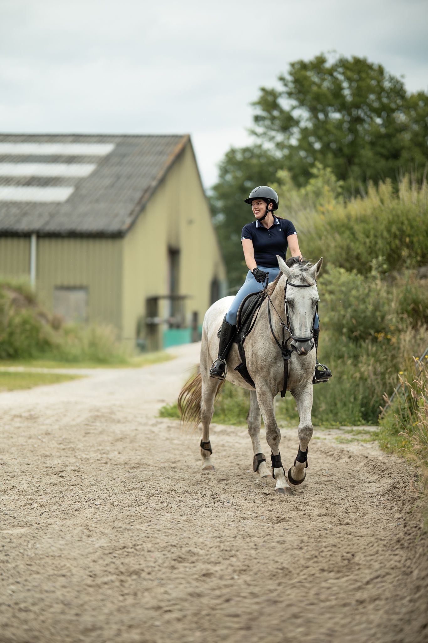 Female equestrian in helmet riding a grey horse along a gravel path near farm buildings.