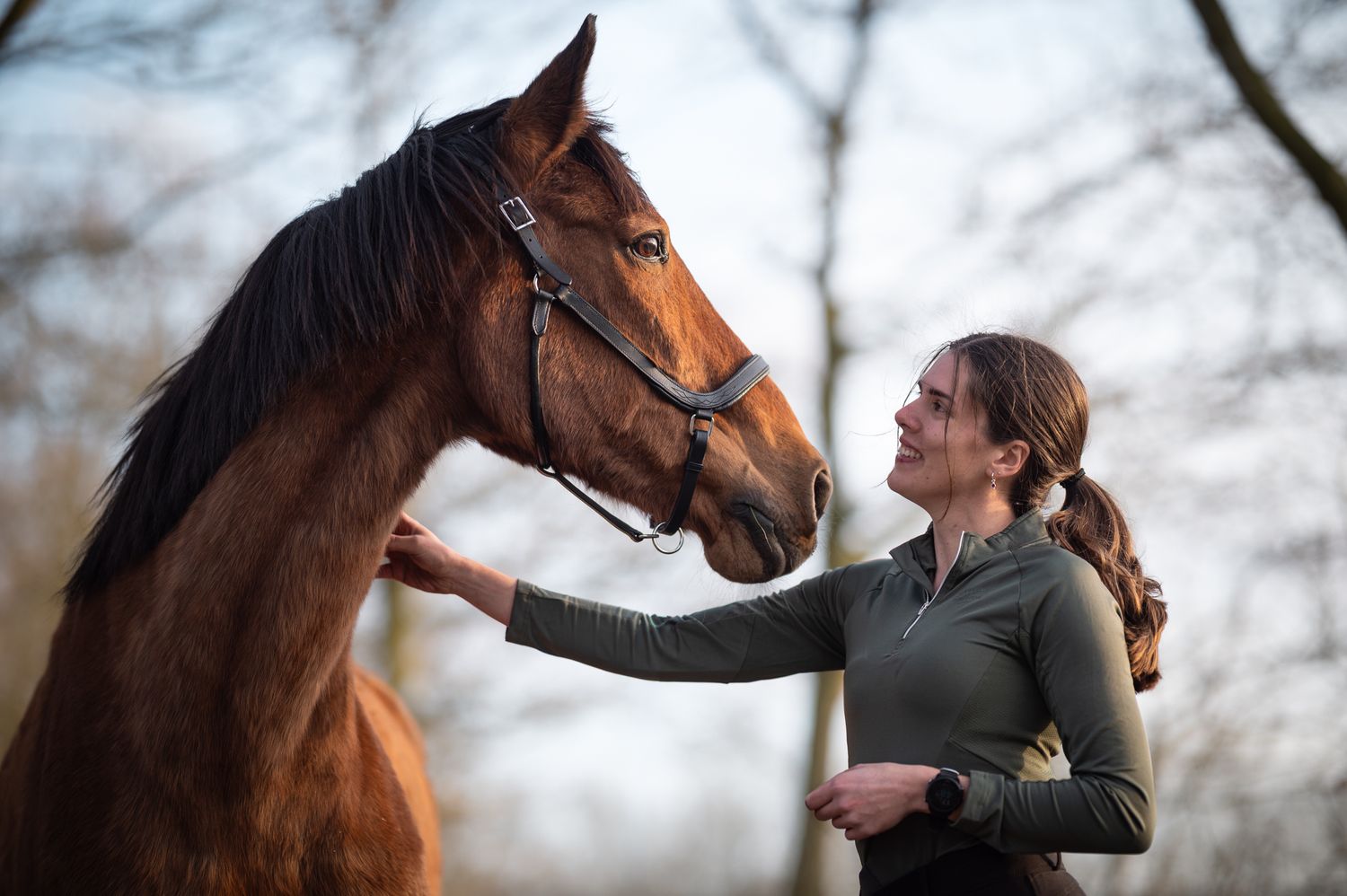 A smiling woman gently stroking the face of a tall chestnut horse outdoors in nature.