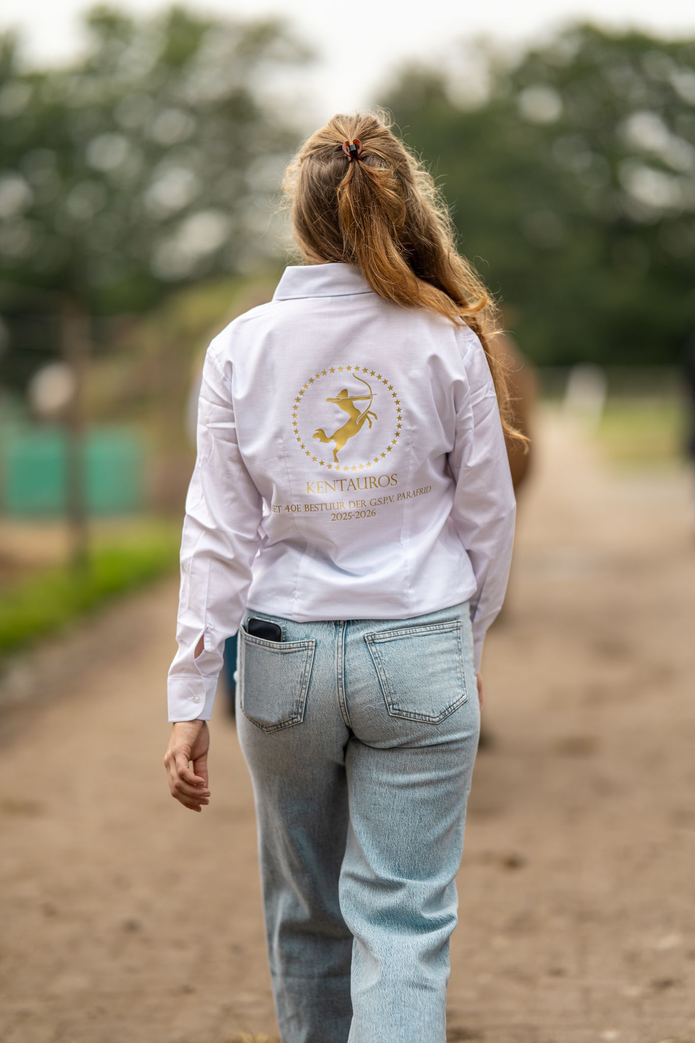 Woman walking away showing white shirt with gold equestrian logo on back.