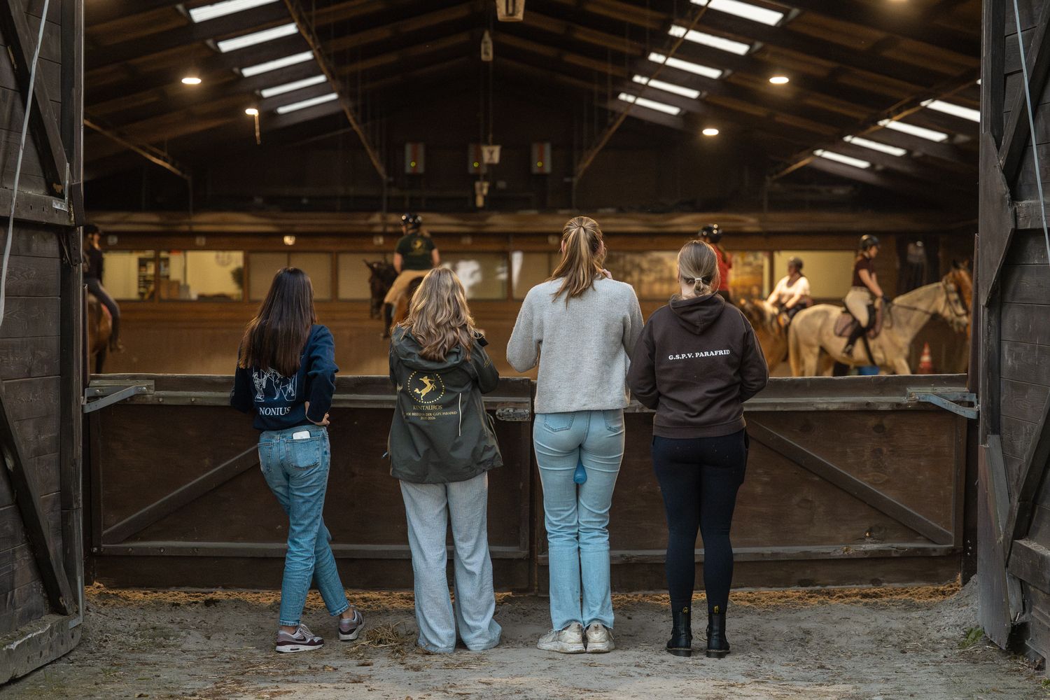Four spectators standing with backs to camera observing an indoor horse riding lesson in a stable arena.