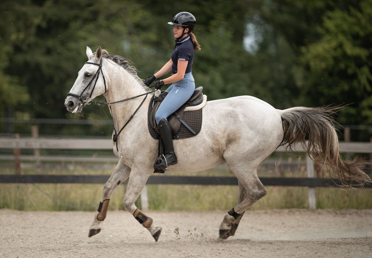 Woman in helmet rides a gray horse at a canter in an outdoor sand arena surrounded by green trees.