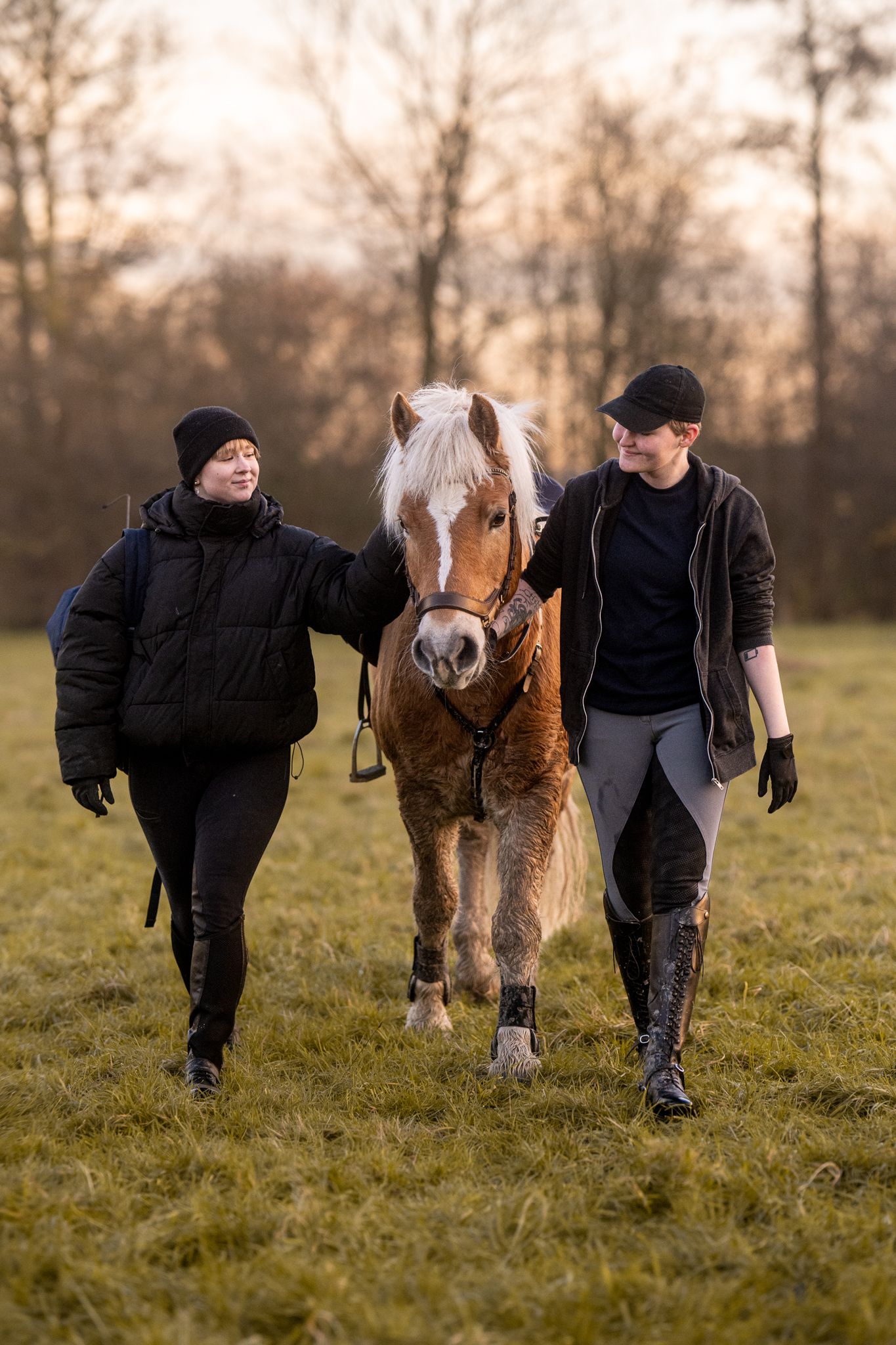 Two women walking a chestnut pony with white mane across a grassy field at sunset.