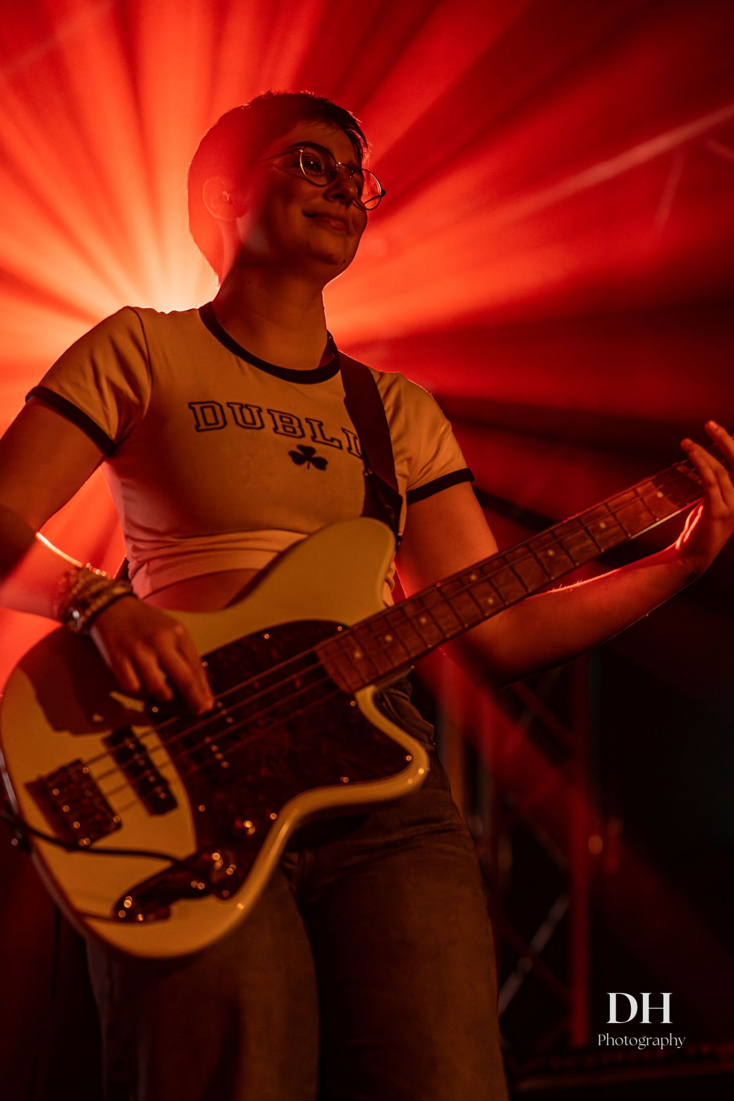 Guitarist performing on stage under dramatic red lighting, wearing a Dublin t-shirt and glasses.