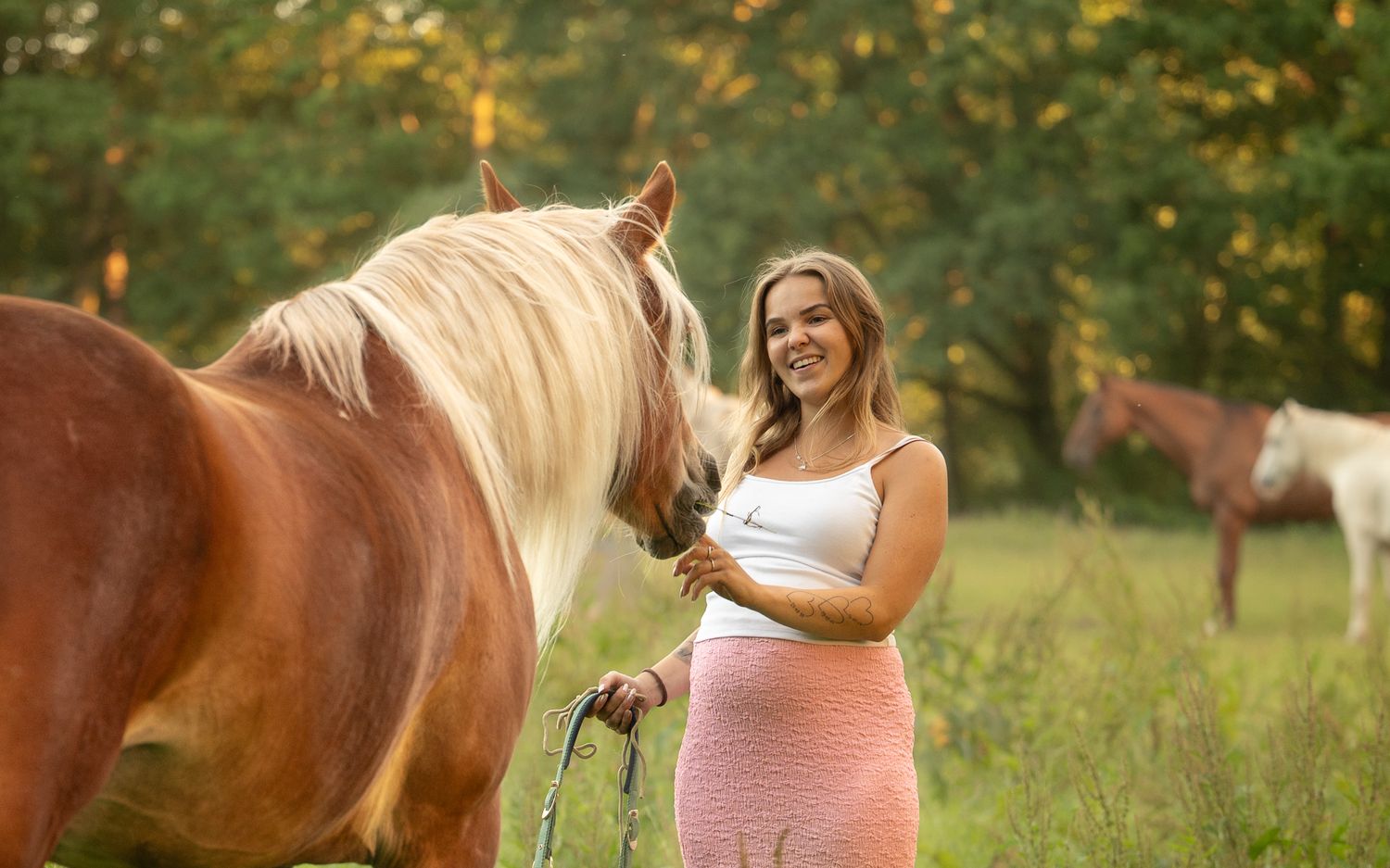 Smiling pregnant woman in pink skirt standing with a palomino horse in a meadow.