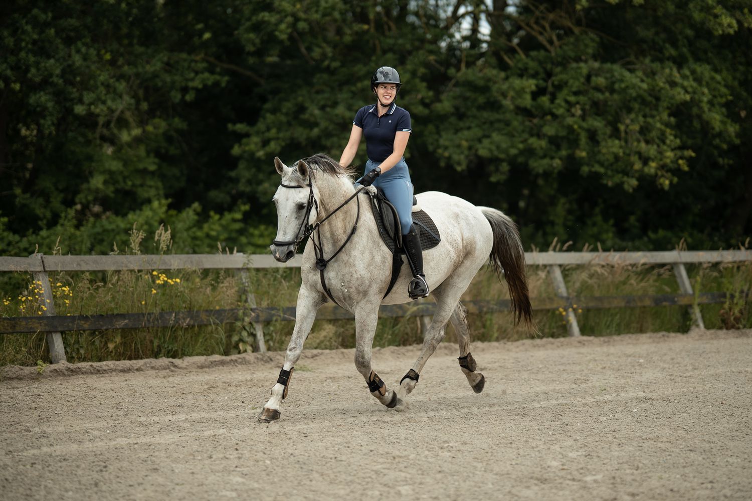 A rider in a helmet and navy top canters a dapple grey horse in an outdoor sand arena surrounded by greenery.