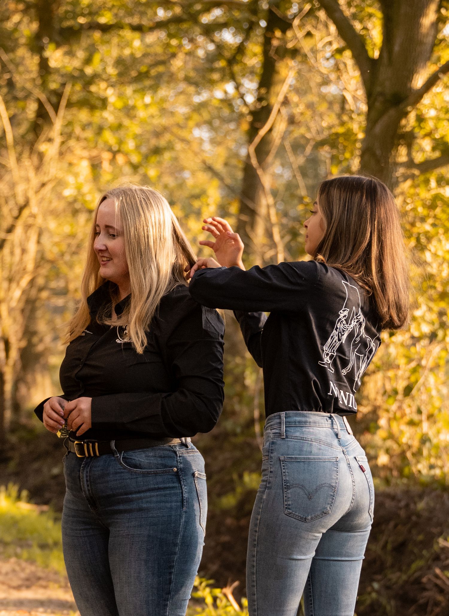 Two young women in black shirts and jeans laughing together in a sunny autumn forest setting.