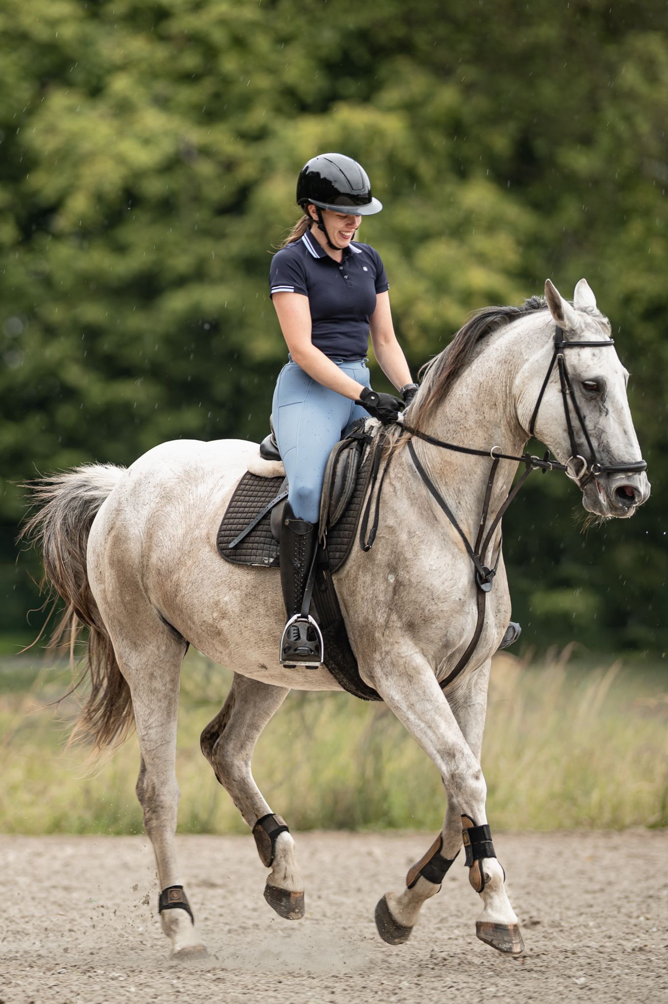 Female equestrian in blue riding gear cantering on a dapple grey horse in an outdoor arena.