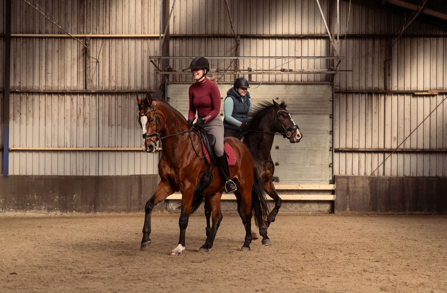 Two riders on dark horses practicing dressage inside an indoor equestrian arena.