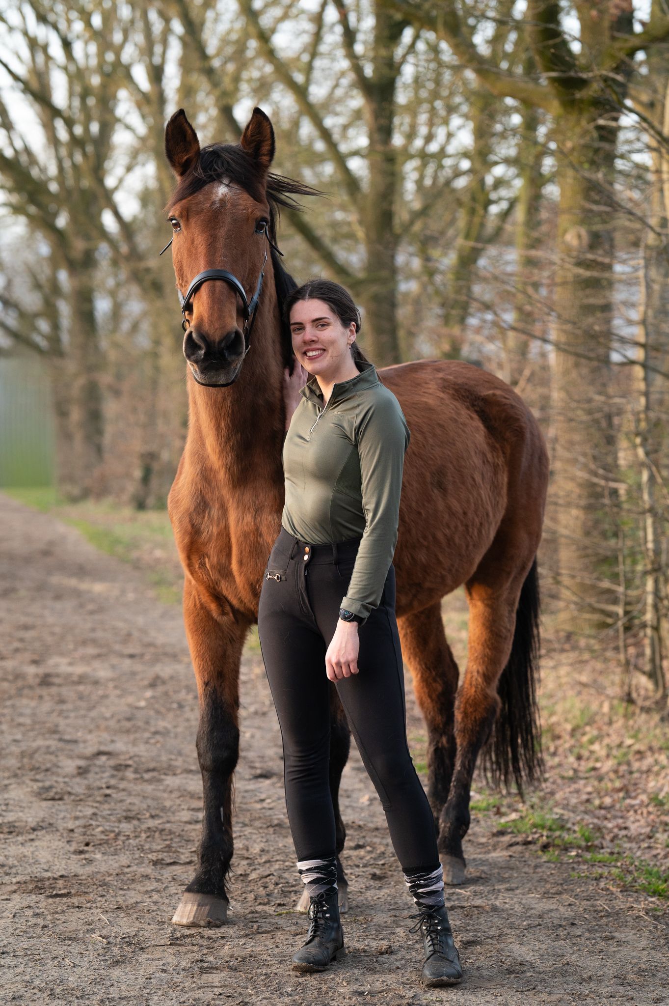 Young woman in equestrian attire smiling beside a bay horse on a tree-lined dirt path.