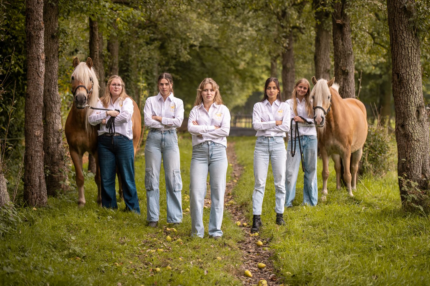 Five women in white shirts and jeans pose confidently with two palomino horses on a tree-lined path.
