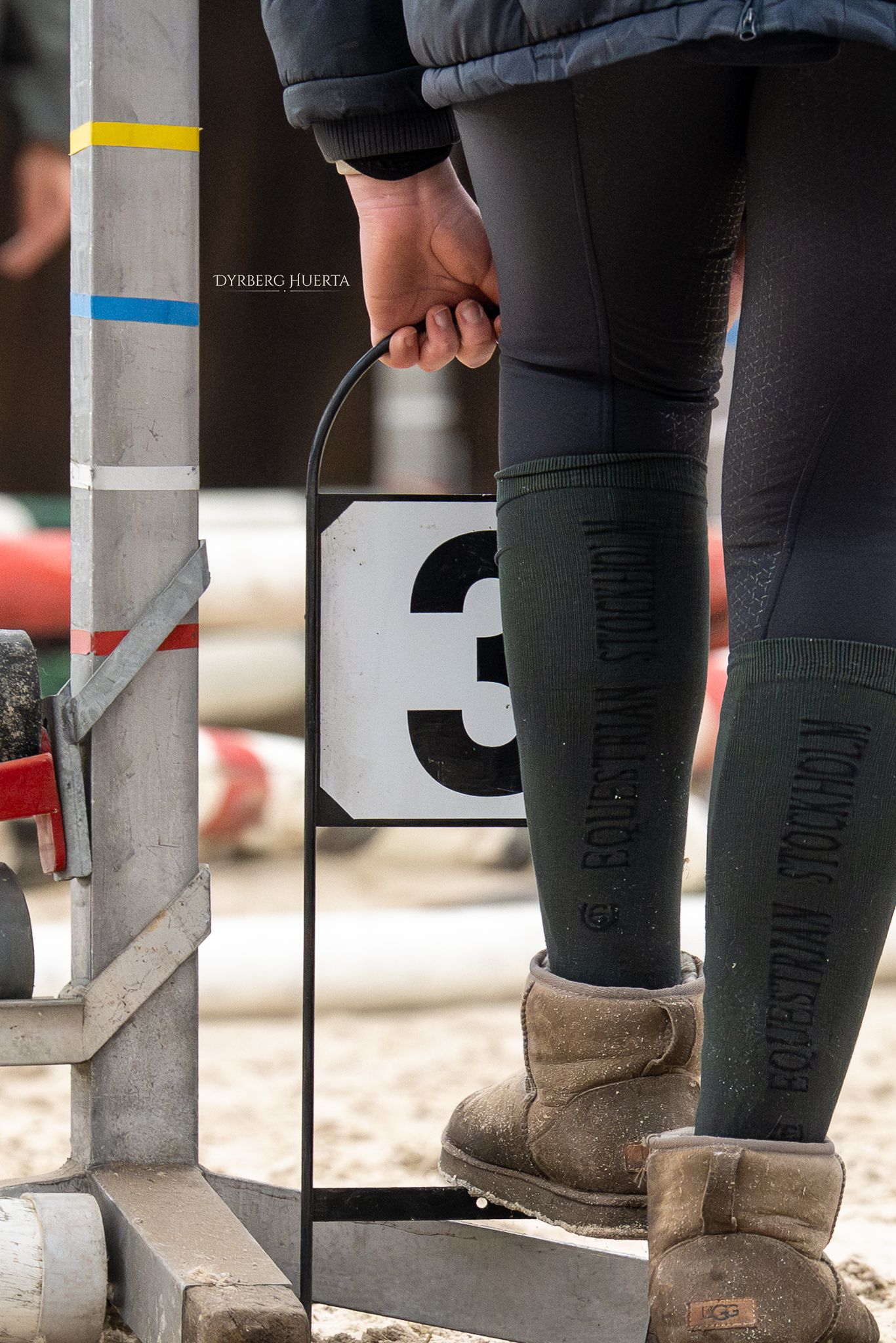 Person holding a number 3 sign at an equestrian show jumping competition, wearing riding boots and breeches.