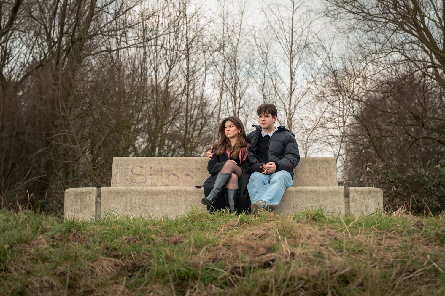 A young couple sits closely together on a stone bench surrounded by bare trees and grass in an outdoor park setting.