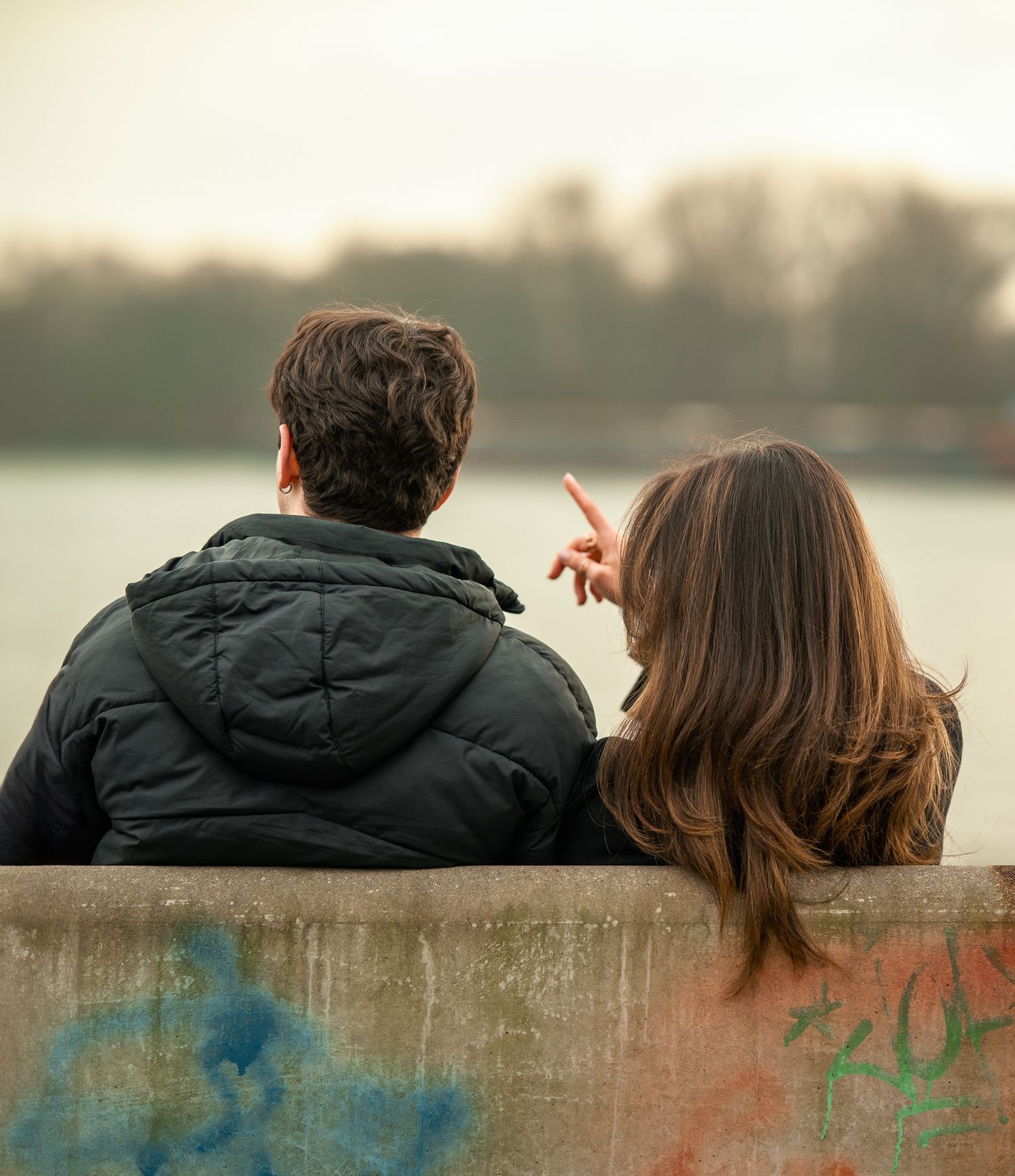 Couple sitting on a graffiti wall holding hands, pointing toward a calm river at dusk.